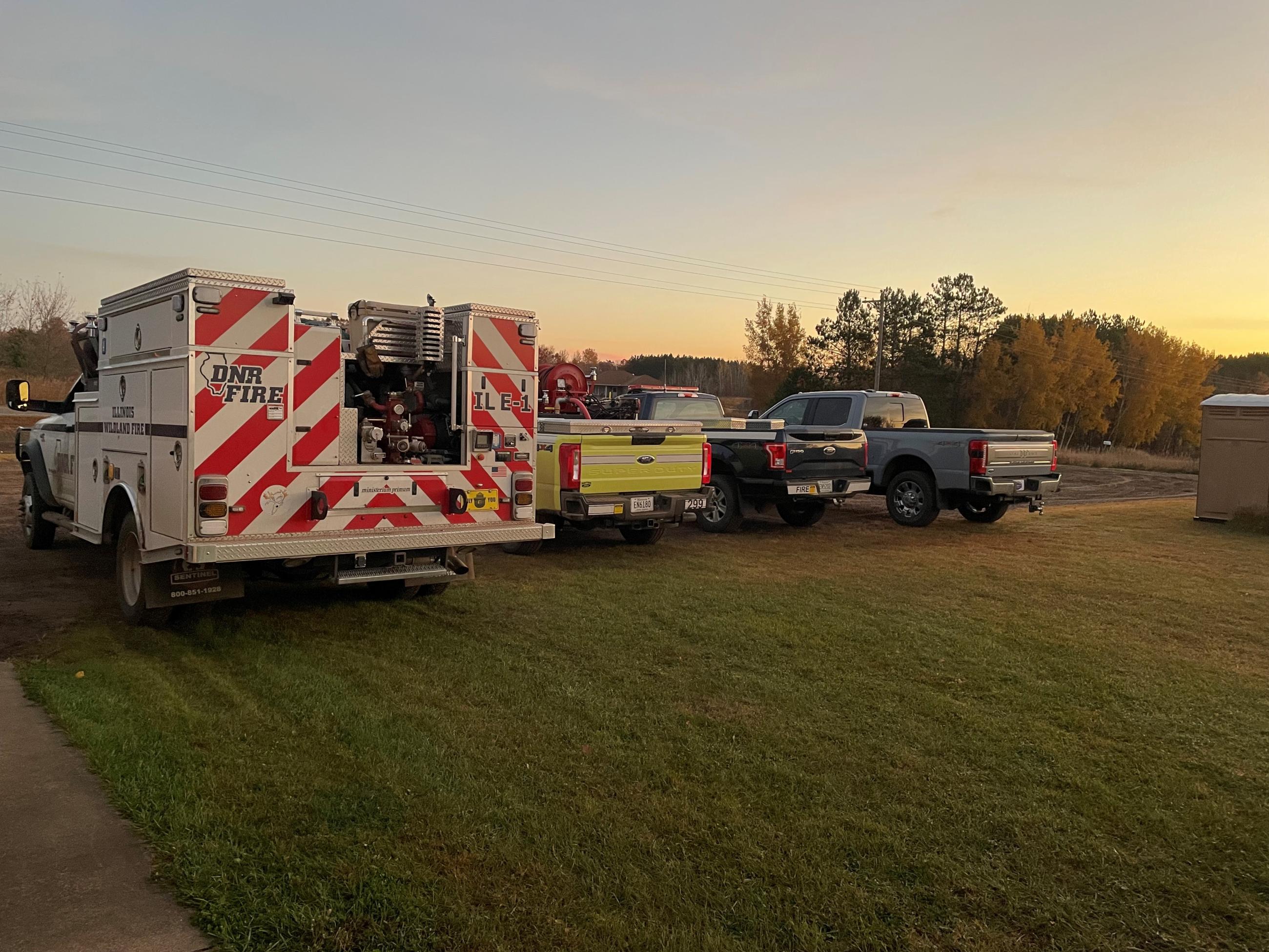 A line of 3 trucks and a fire engine parked on grass. 