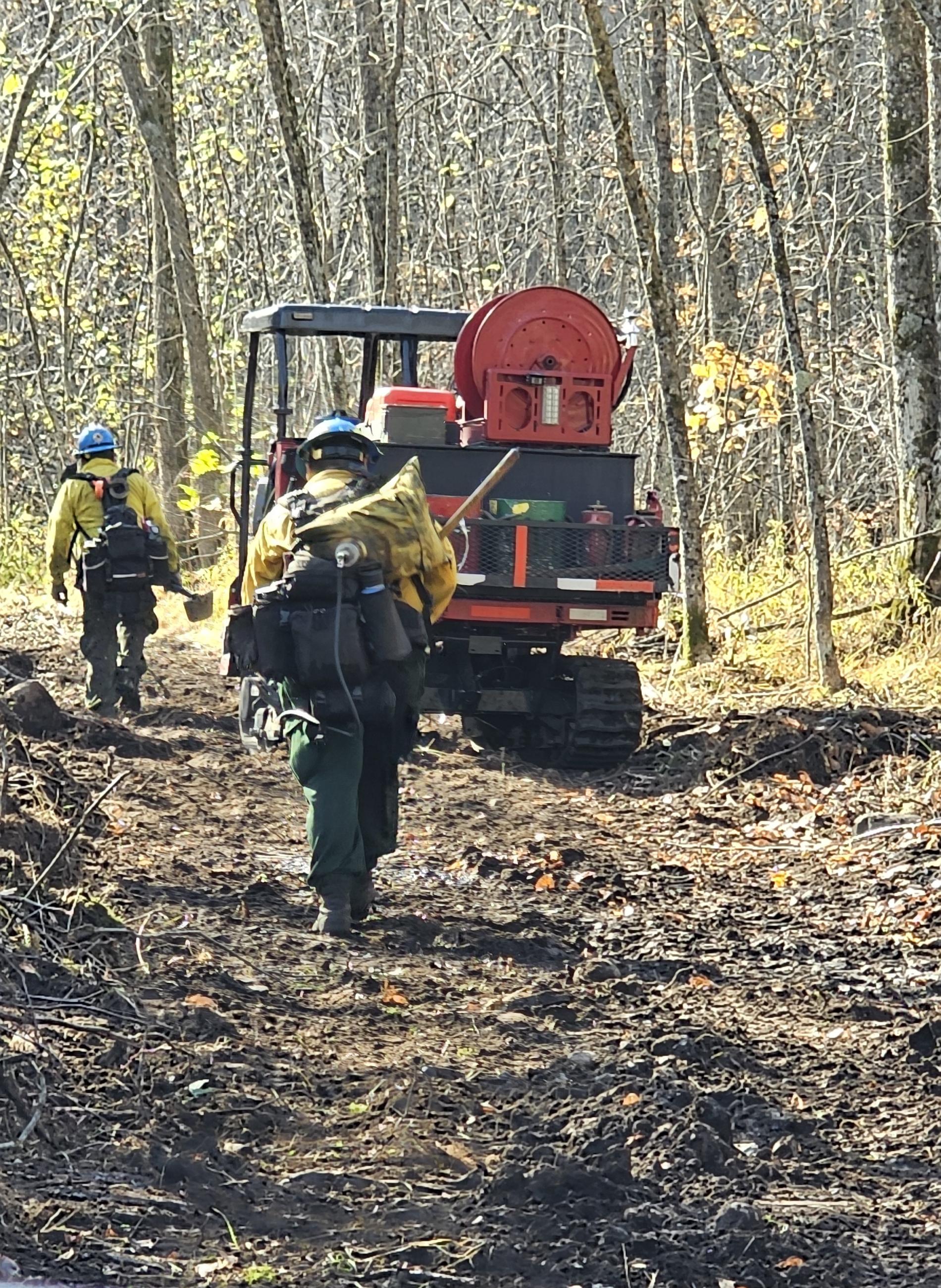 Two firefighters walk by an ATV on a dozer line in firefighting gear. 