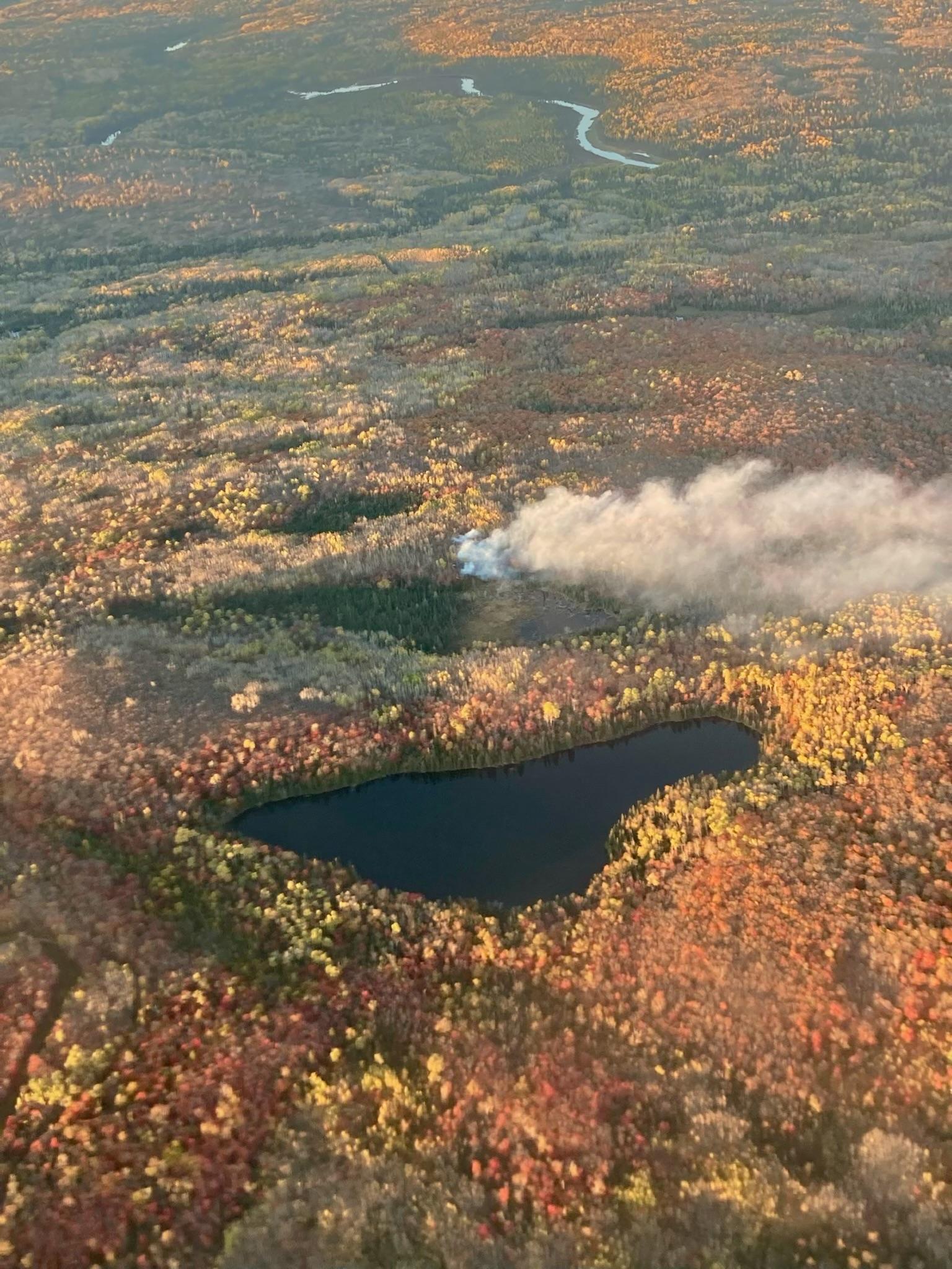 Aerial photo of white smoke rising from the forest with fall colors on the trees and Bogus Lake in the foreground