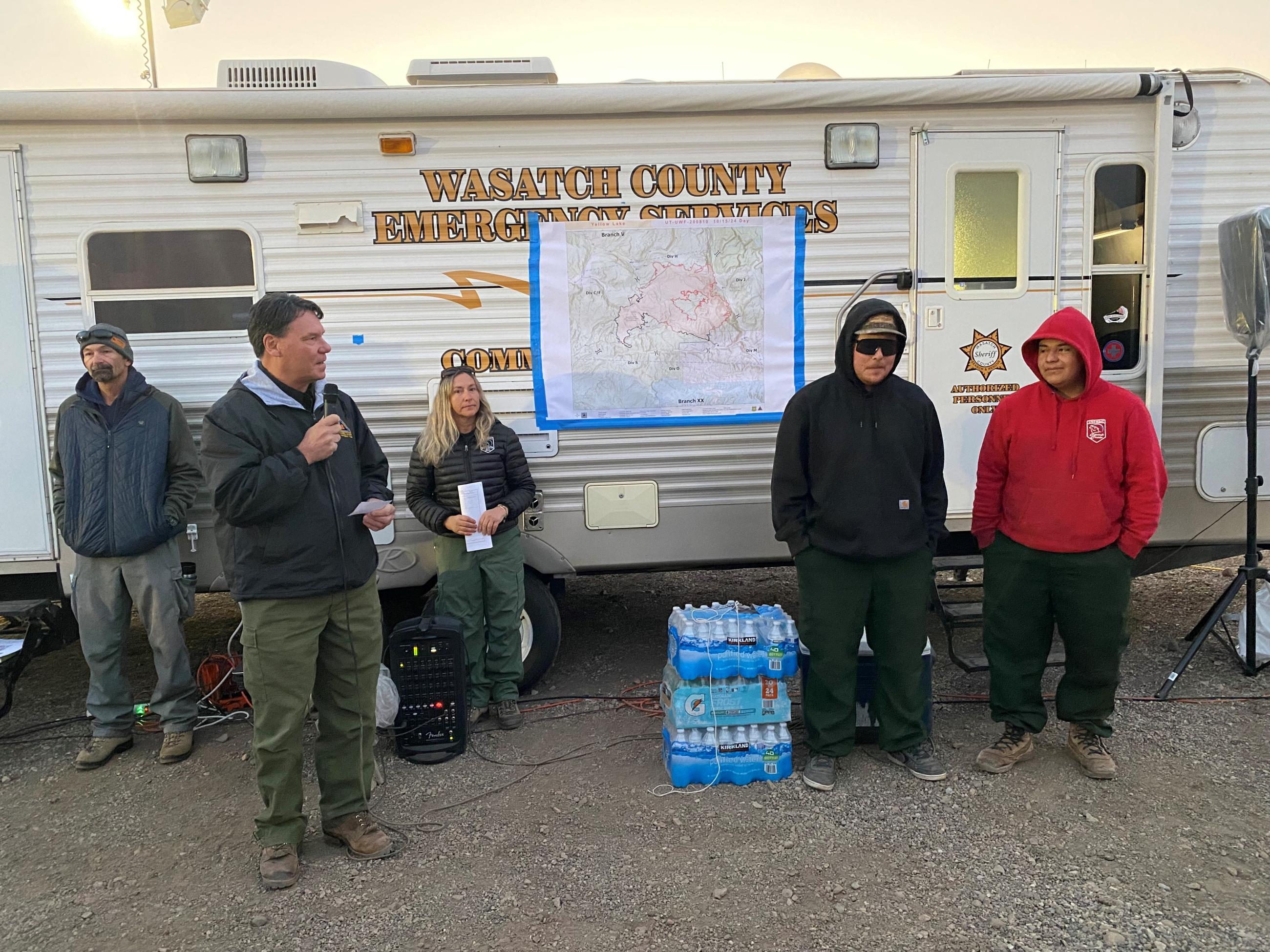 Incident Commander speaks into a microphone and stands among four other individuals outside.