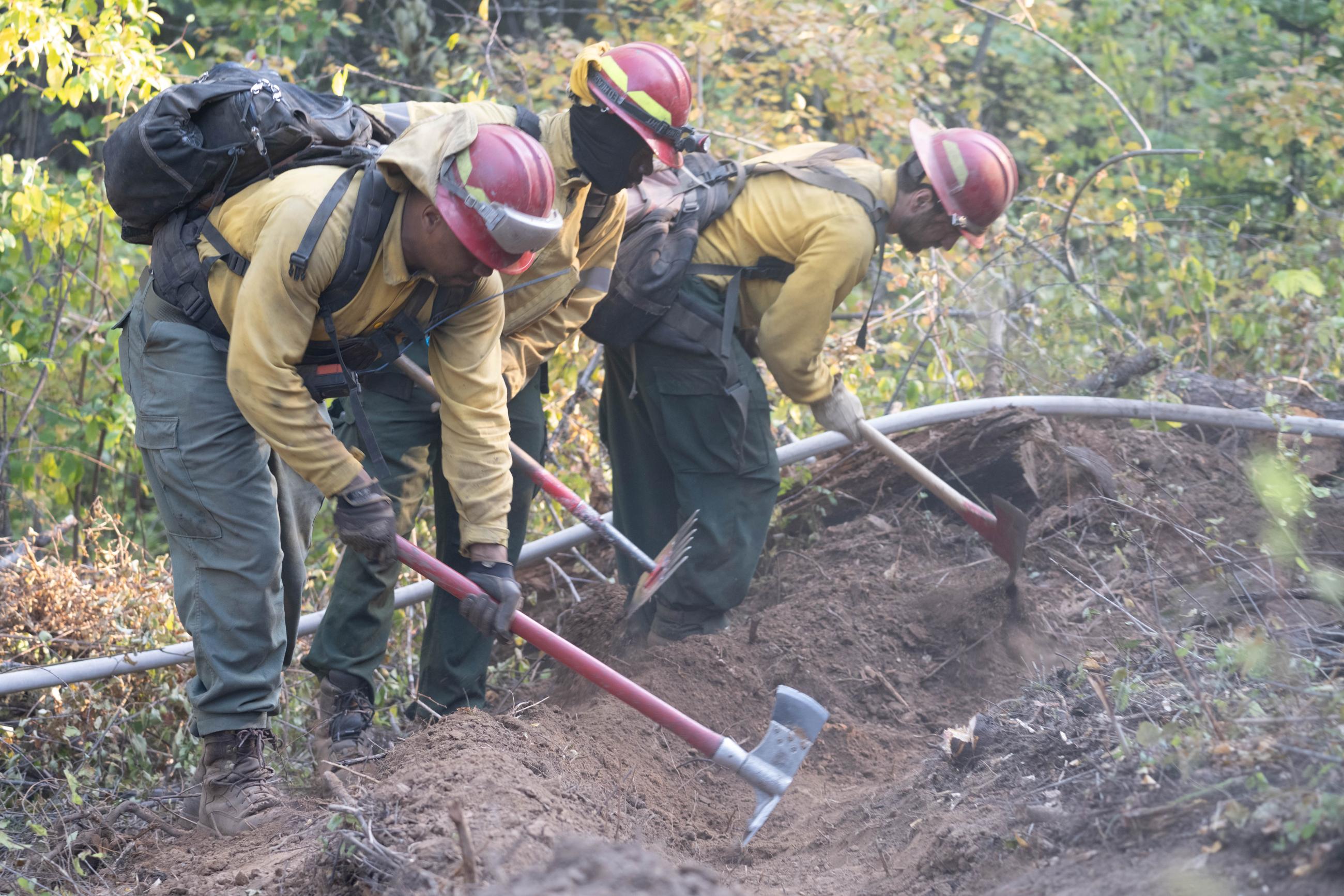 Three firefighters swinging tools into soil