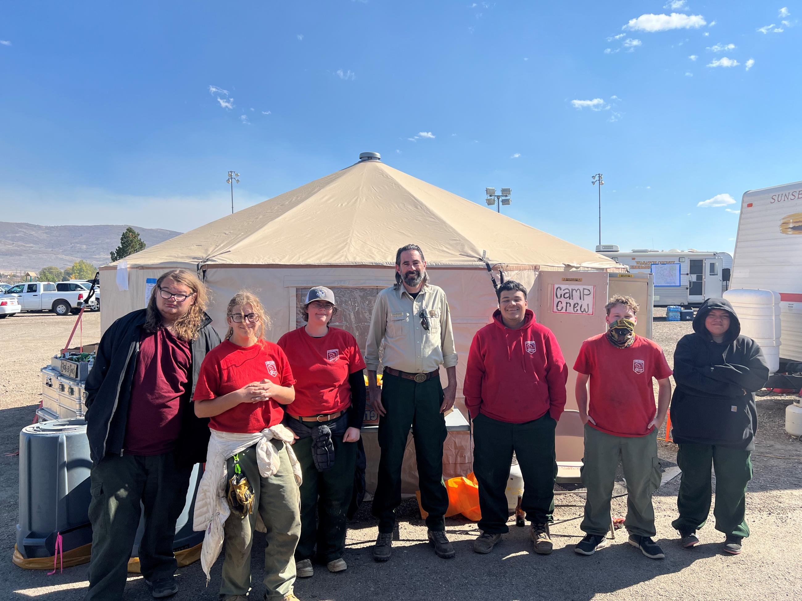 Group stands outside under blue skies in front of a fire camp yurt.