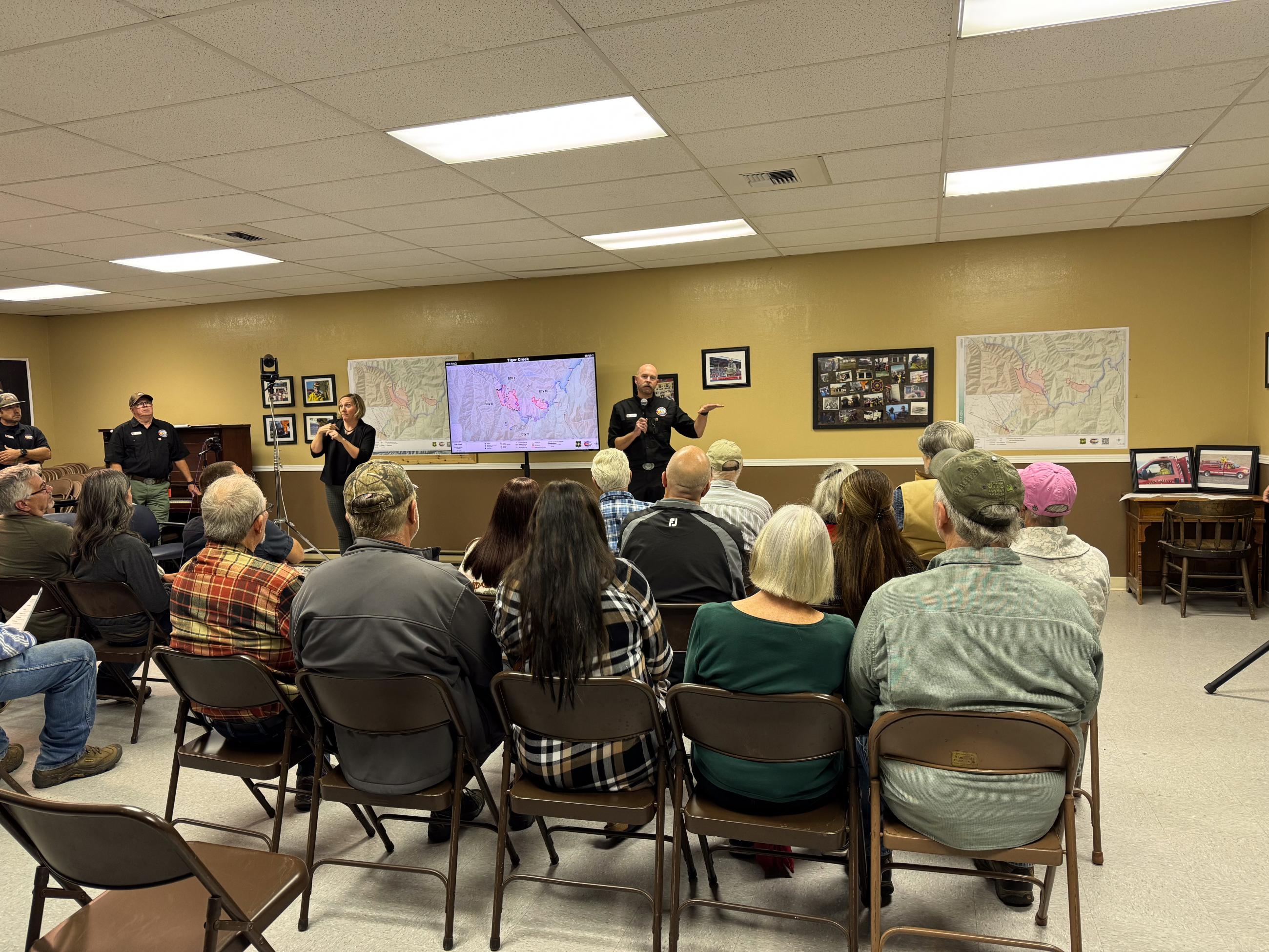 Large group sitting facing a sign language interpreter and several maps on the wall.