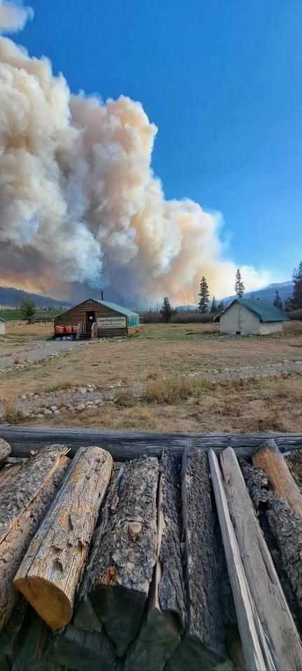 A large column of smoke is seen in the distance against a blue sky with a cabin in the middle distance and stacked firewood in the foreground.