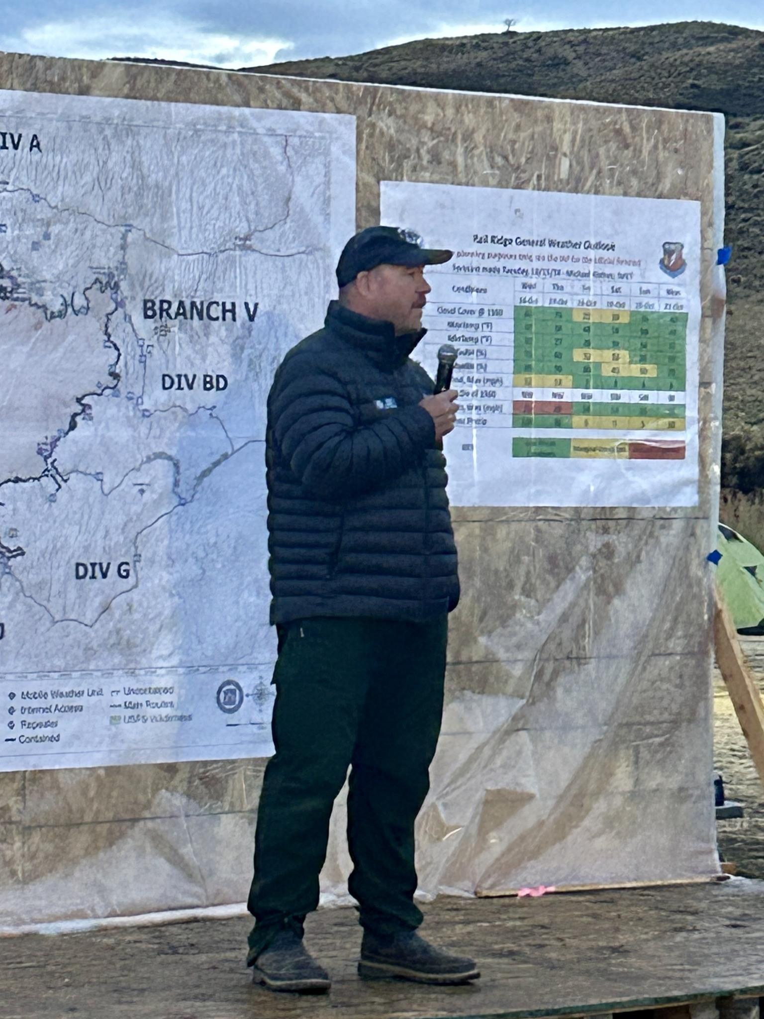 John Szulc is pictured on stage providing information to fire personnel with maps in the background