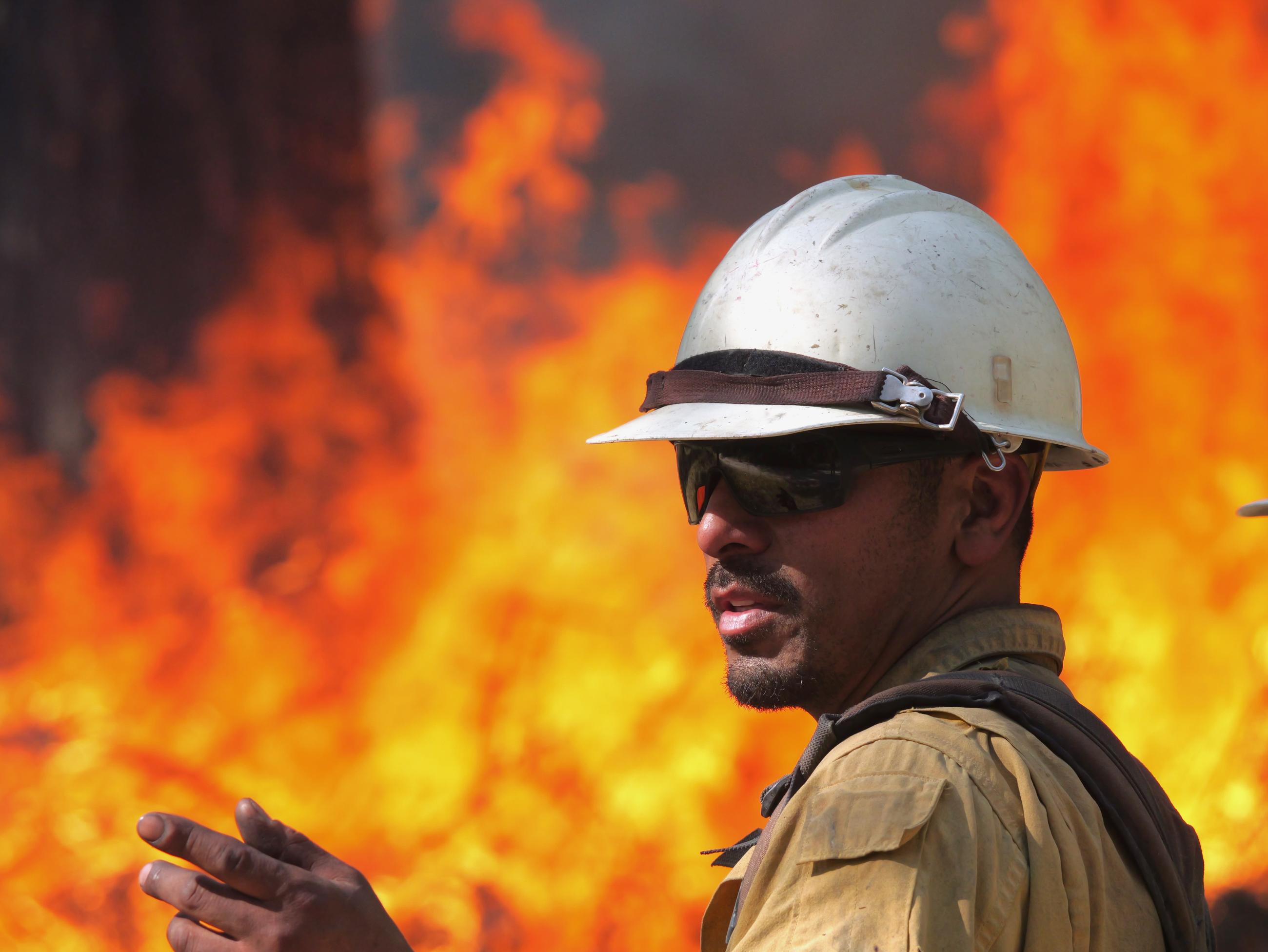 A male firefighter in a yellow shirt and white helmet points, framed by flames in the distance.