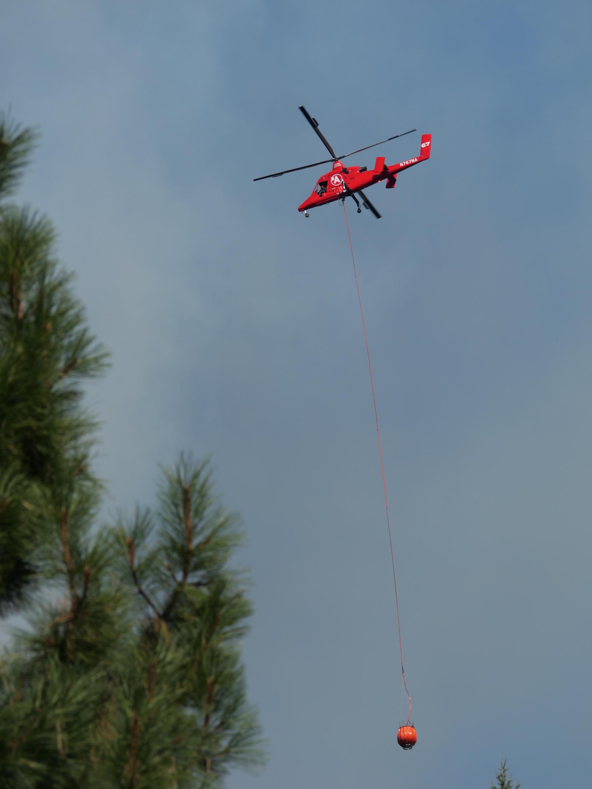 A red KMAX helicopter trailing an orange bucket full of water passes over trees on its way to cool a hotspot on October 2, 2024.