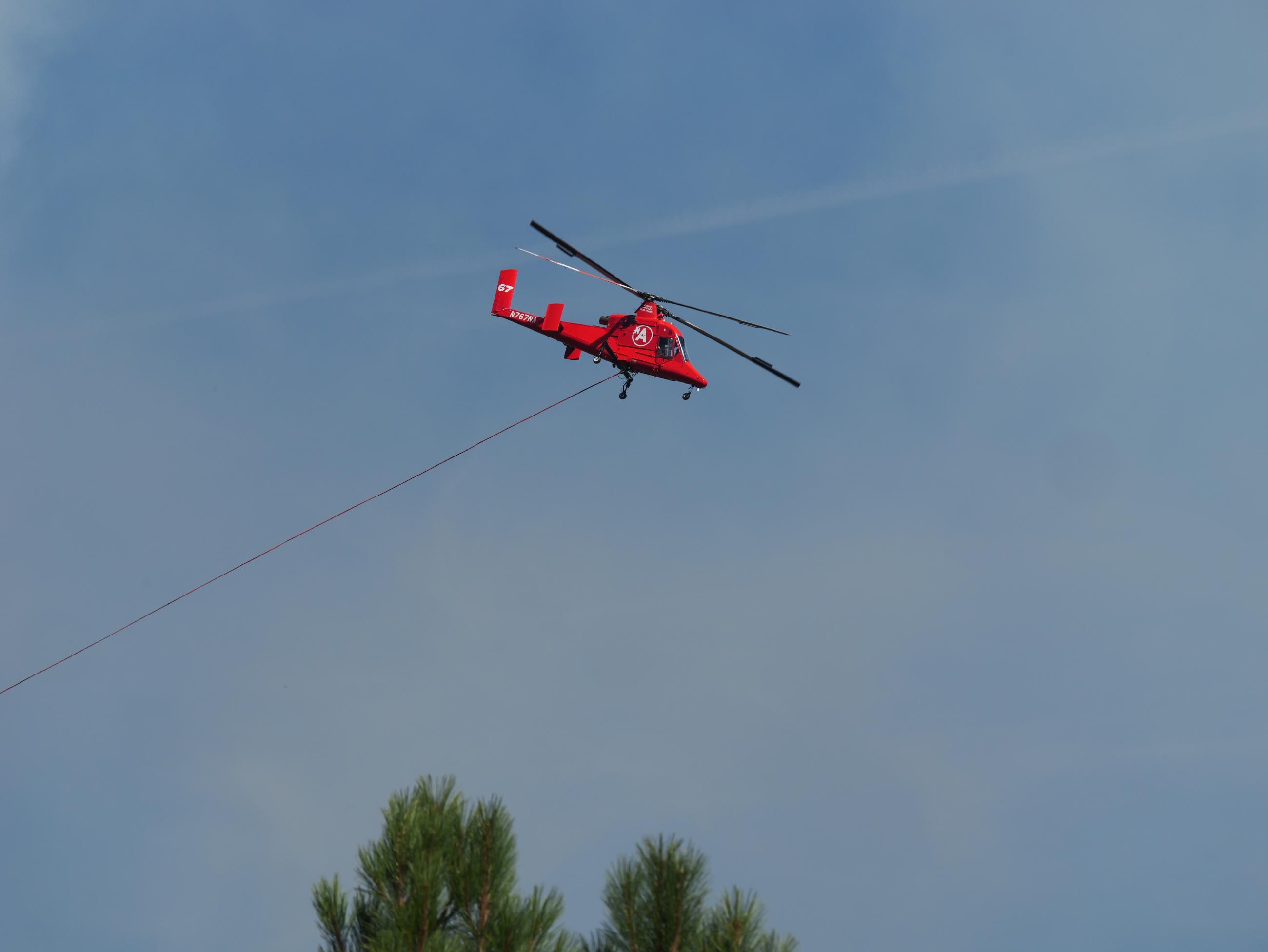 A red helicopter trailing a cable passes through a mostly blue sky above trees.