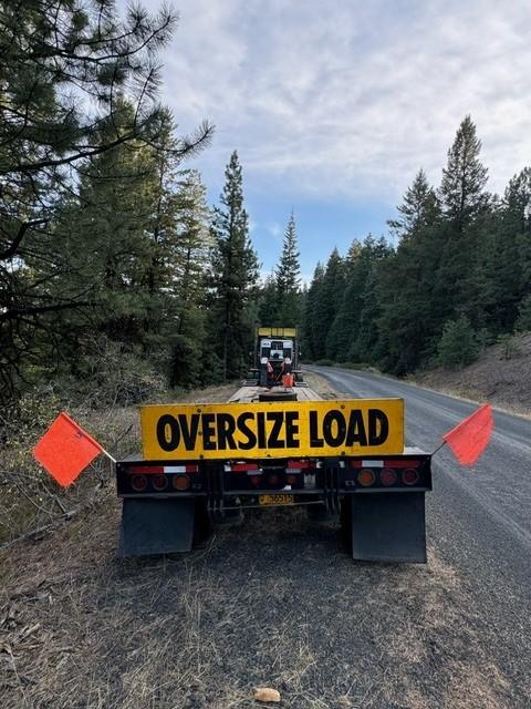 A semi trailer used to haul heavy machinery is shown on a narrow forest service road