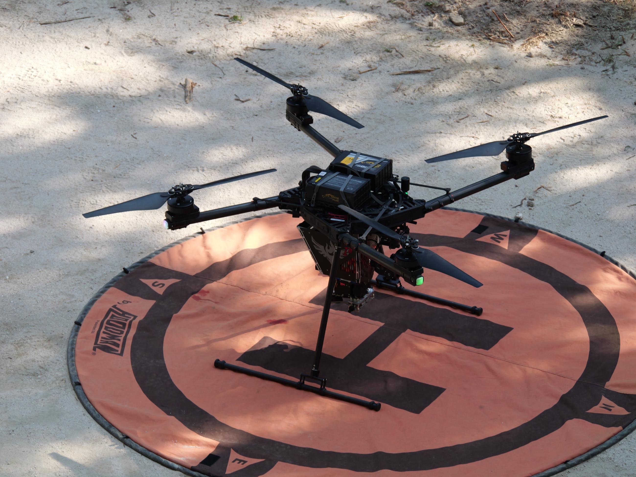 A drone with four rotors sits on an orange pad with the letter "H" within a black circle..