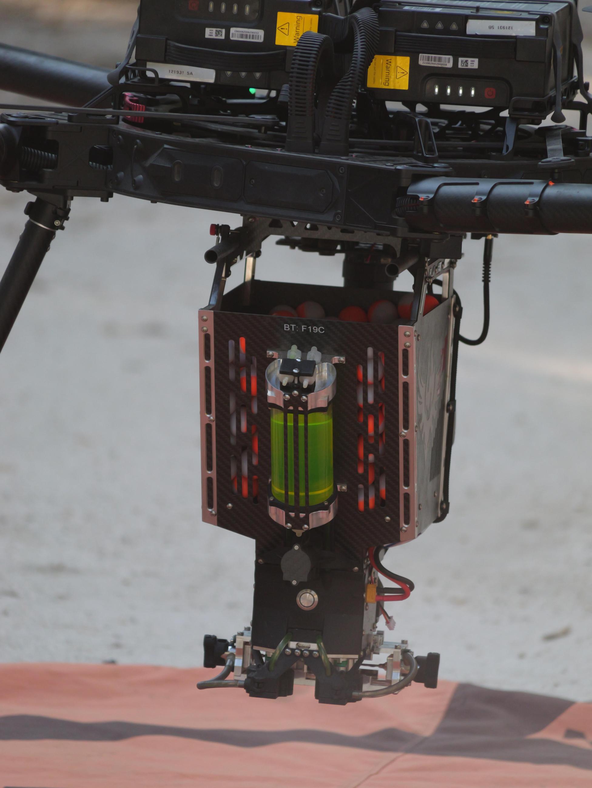 A mechanism attached to a drone is shown, close up. Orange and white plastic balls and a container of yellow-green anti-freeze are part of the mechanism.