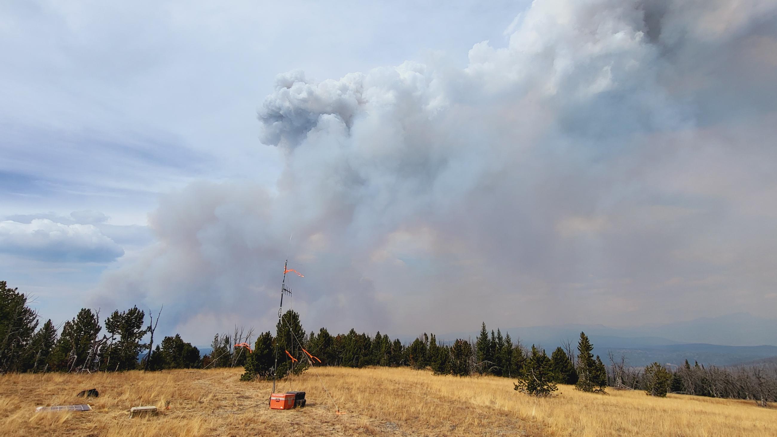 A radio antennae, solar panel, and equipment set in a clearing on top of Fish Lake Mountain. Smoke column rising in the background on a cloudy day  