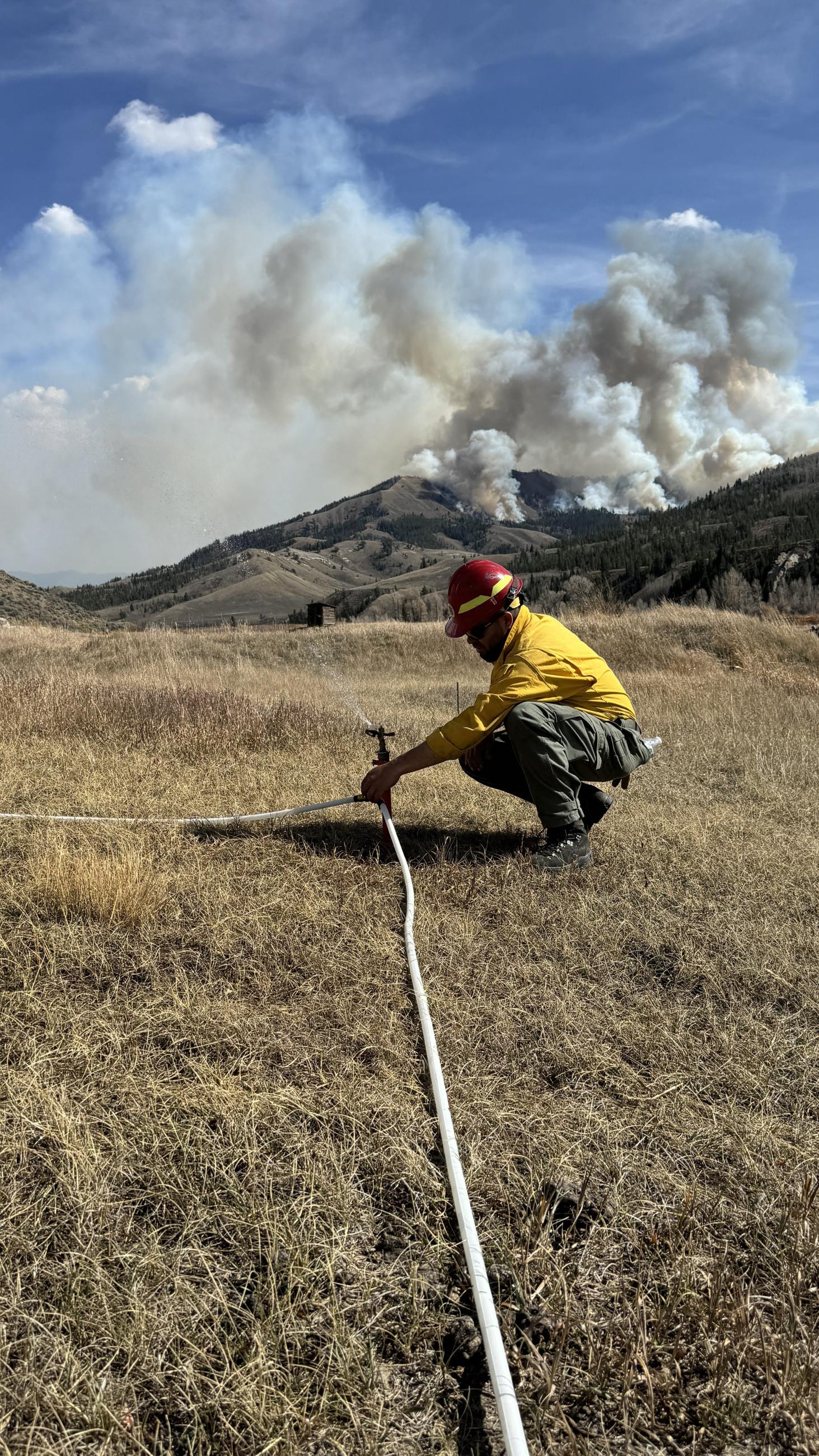 a smoke column rises on a hillside behind a firefighter that is checking a yard sprinkler connected to a white hose