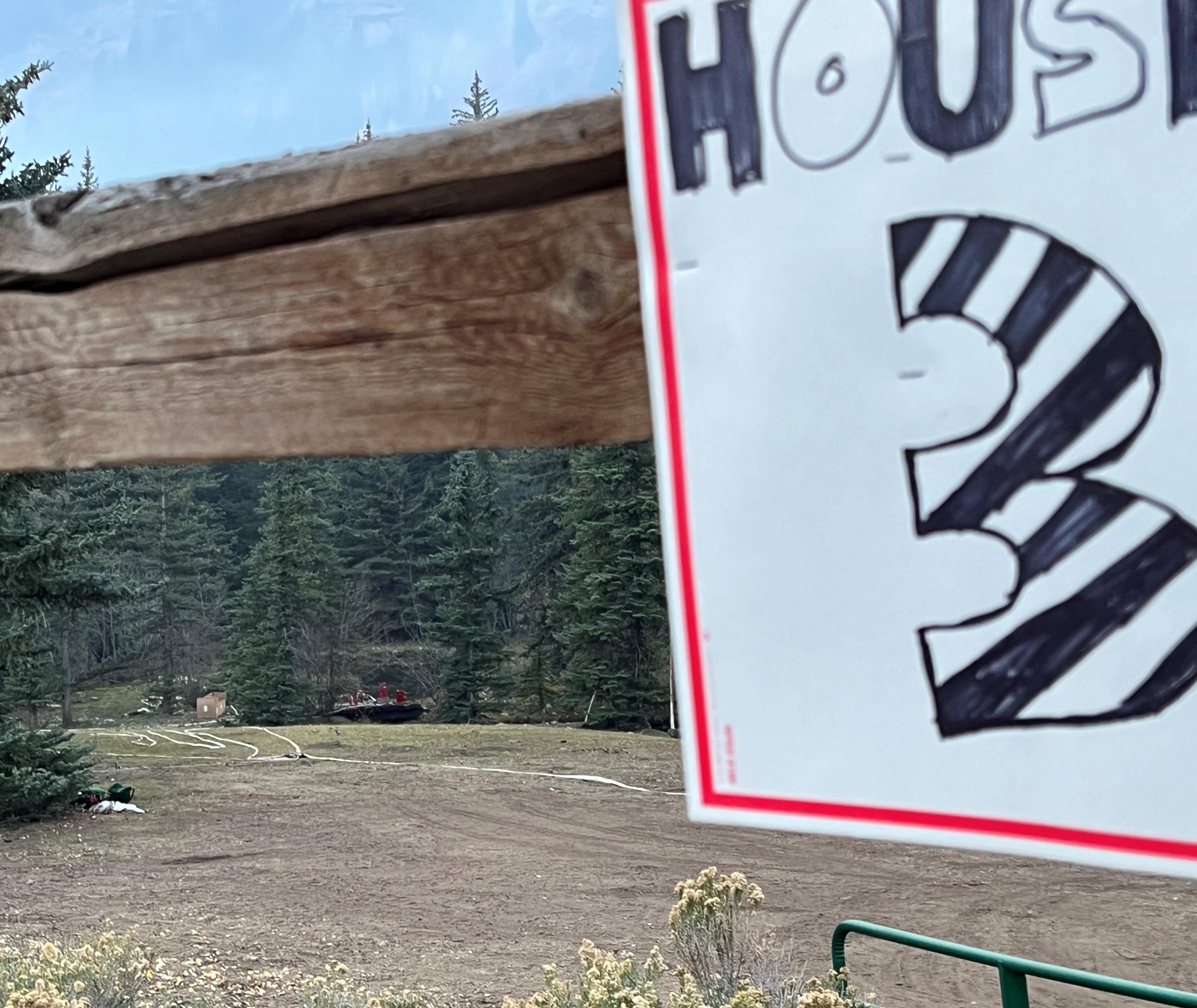 Foreground sign has "House 3" written in black and white with forest and house visible in the far background,