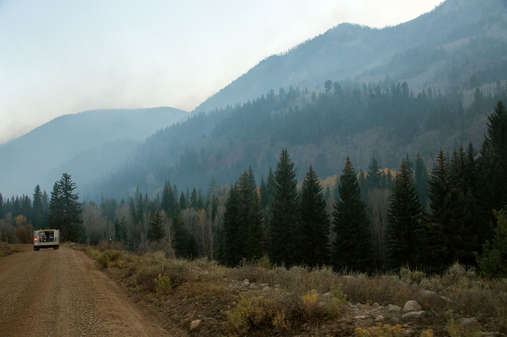 A dirt road sits on the left side of the frame with a fire engine in the distance and pine trees and mountains to the right side of the frame.