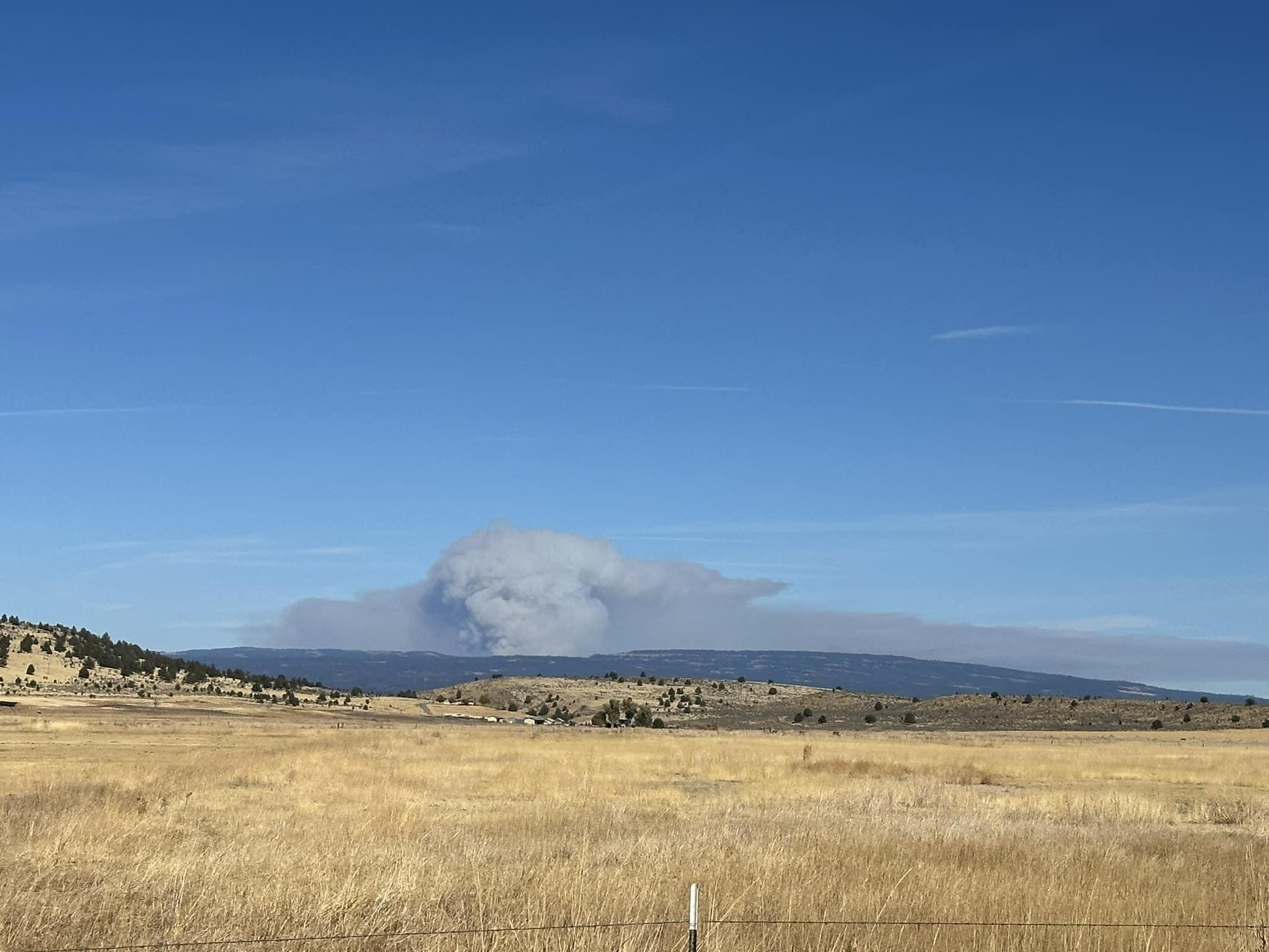 A smoke column is visible past rolling hills and wheat fields