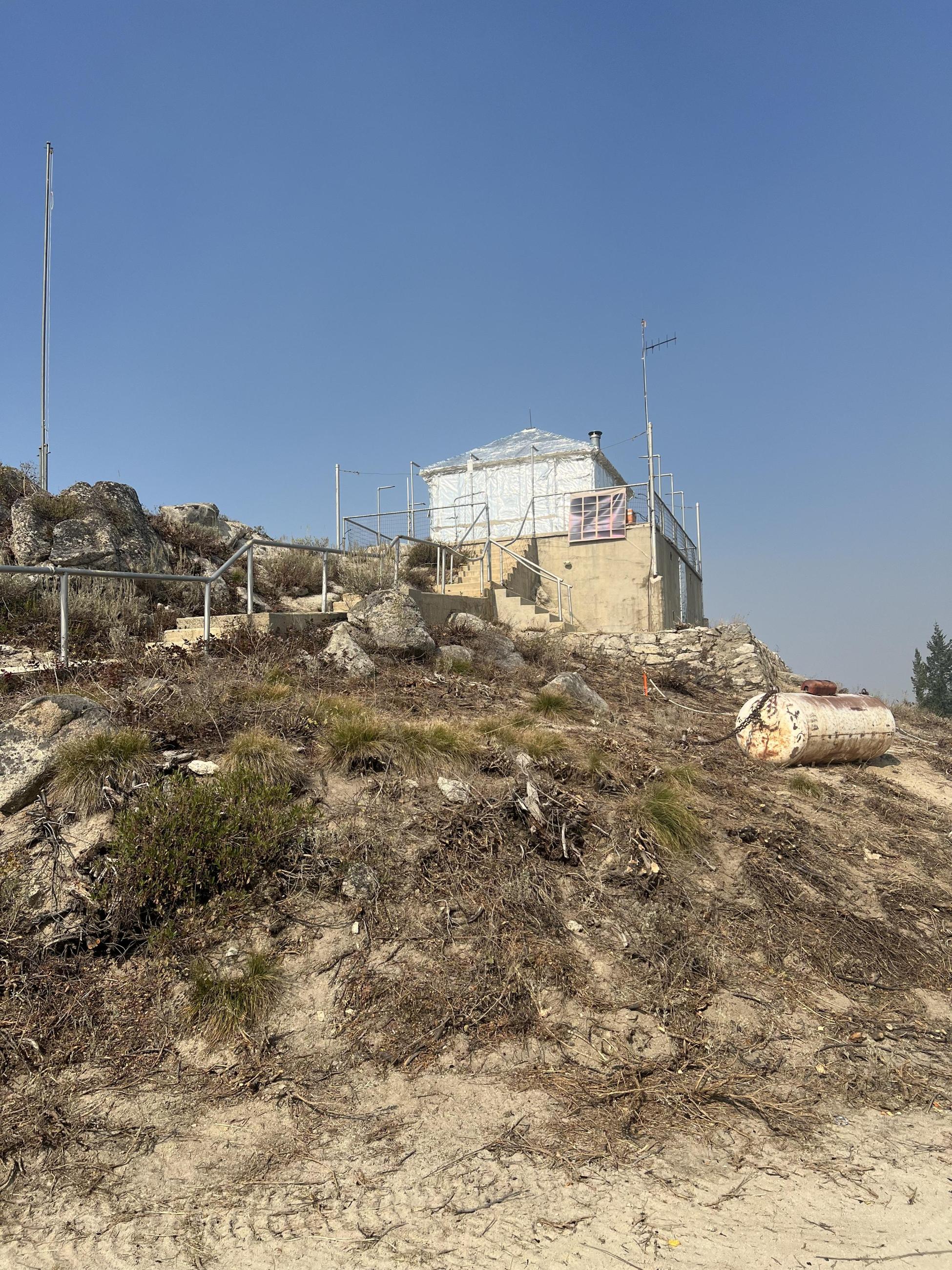 A forest lookout tower, covered in fire resistant foil, is seen against a blue sky.