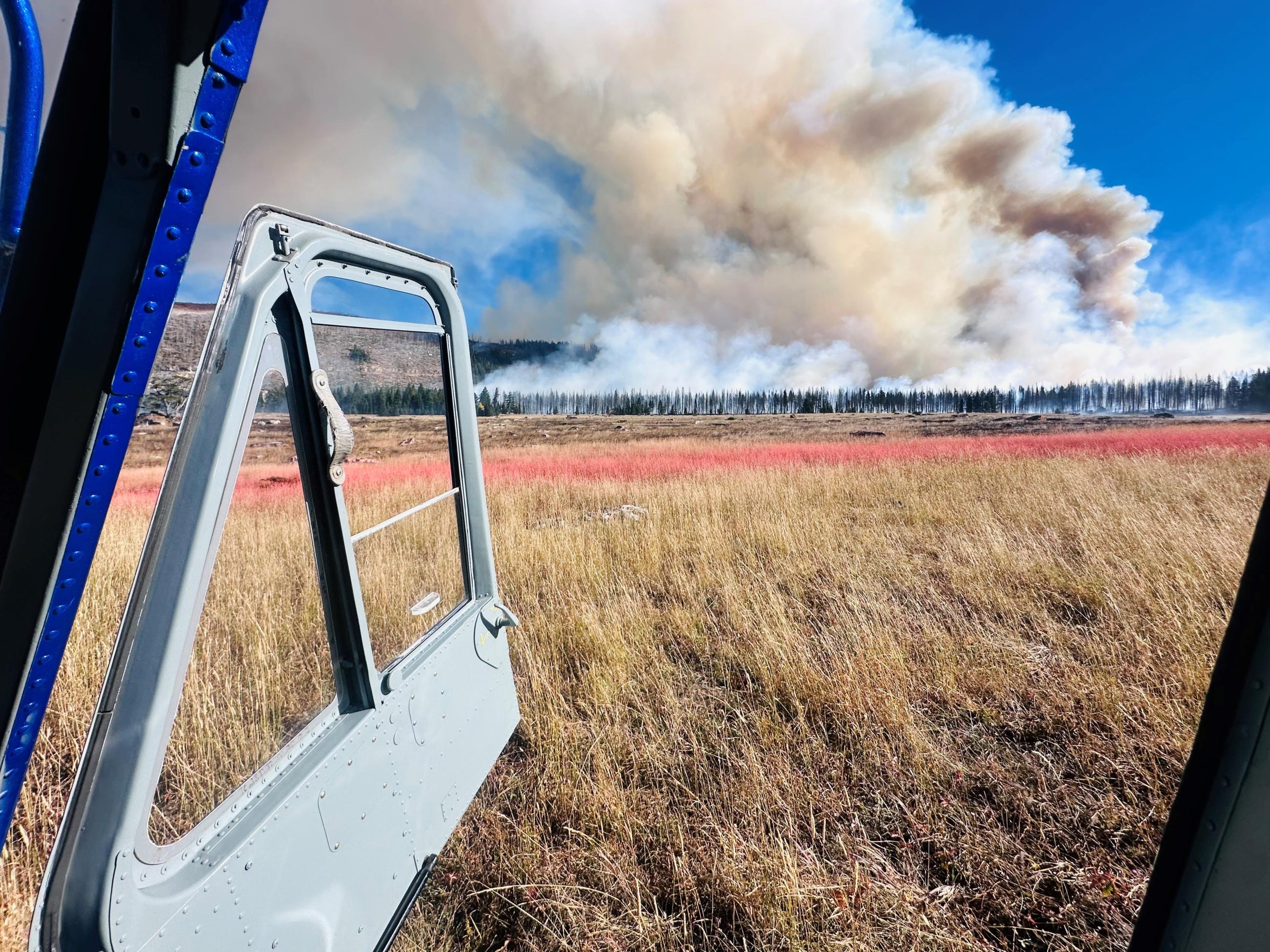 This image shows the view from inside a vehicle parked with its door open. There is a golden field with a line of red retardant coloring the grass vertically right before a line of burning timber. Smoke is pouring from the timber and a column is flowing from the trees to the top left of the image.