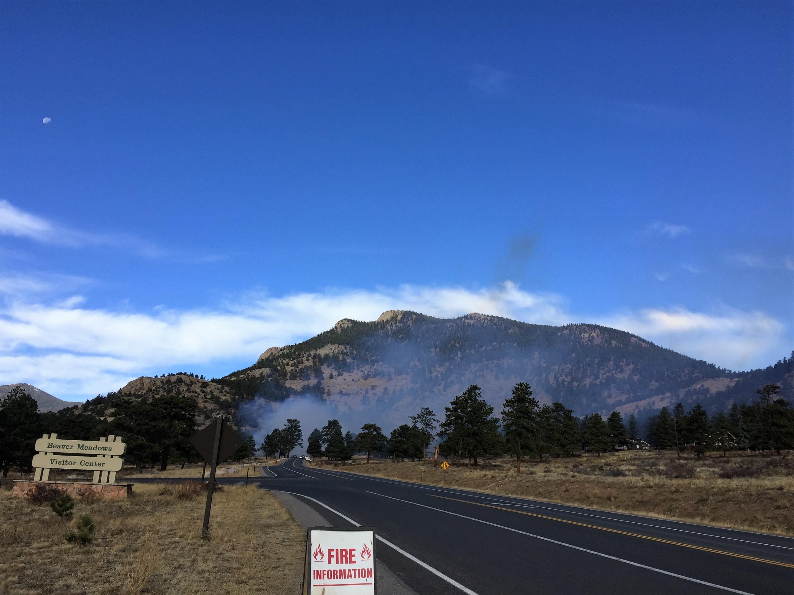 Smoke rises from alongside a roadway during the 2018 Beaver Meadows prescribed fire in Rocky Mountain National Park; two signs are in the foreground along the roadway. One reads 'fire information,' the other is a large sign that says Beaver Meadows Visitor Center. A mountain with trees is in the background and smoke is rising from the trees in front of the mountain. 