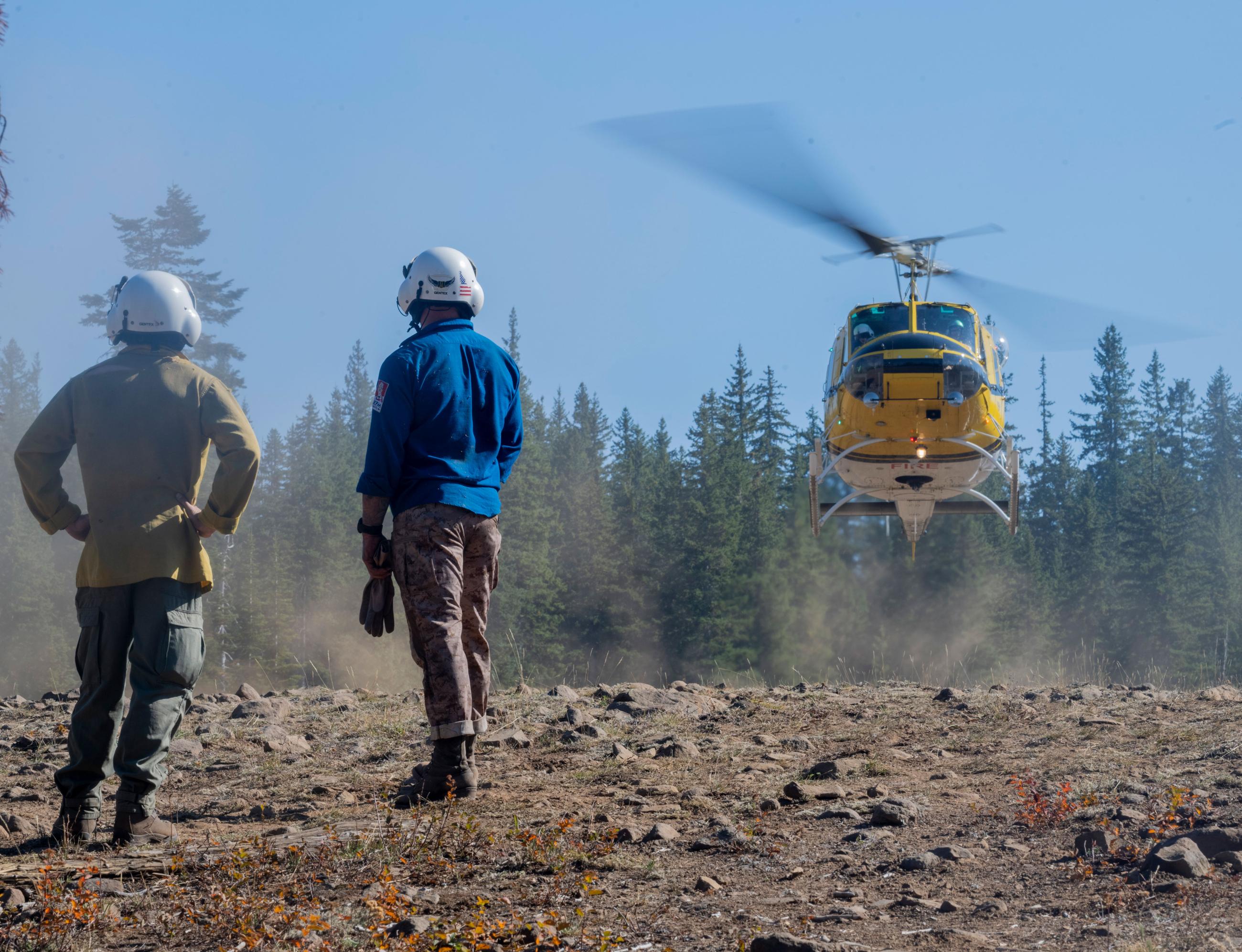 Two people wearing white helmets stand in the foreground with their backs turned to the camera as they watch a helicopter take off from the ground.