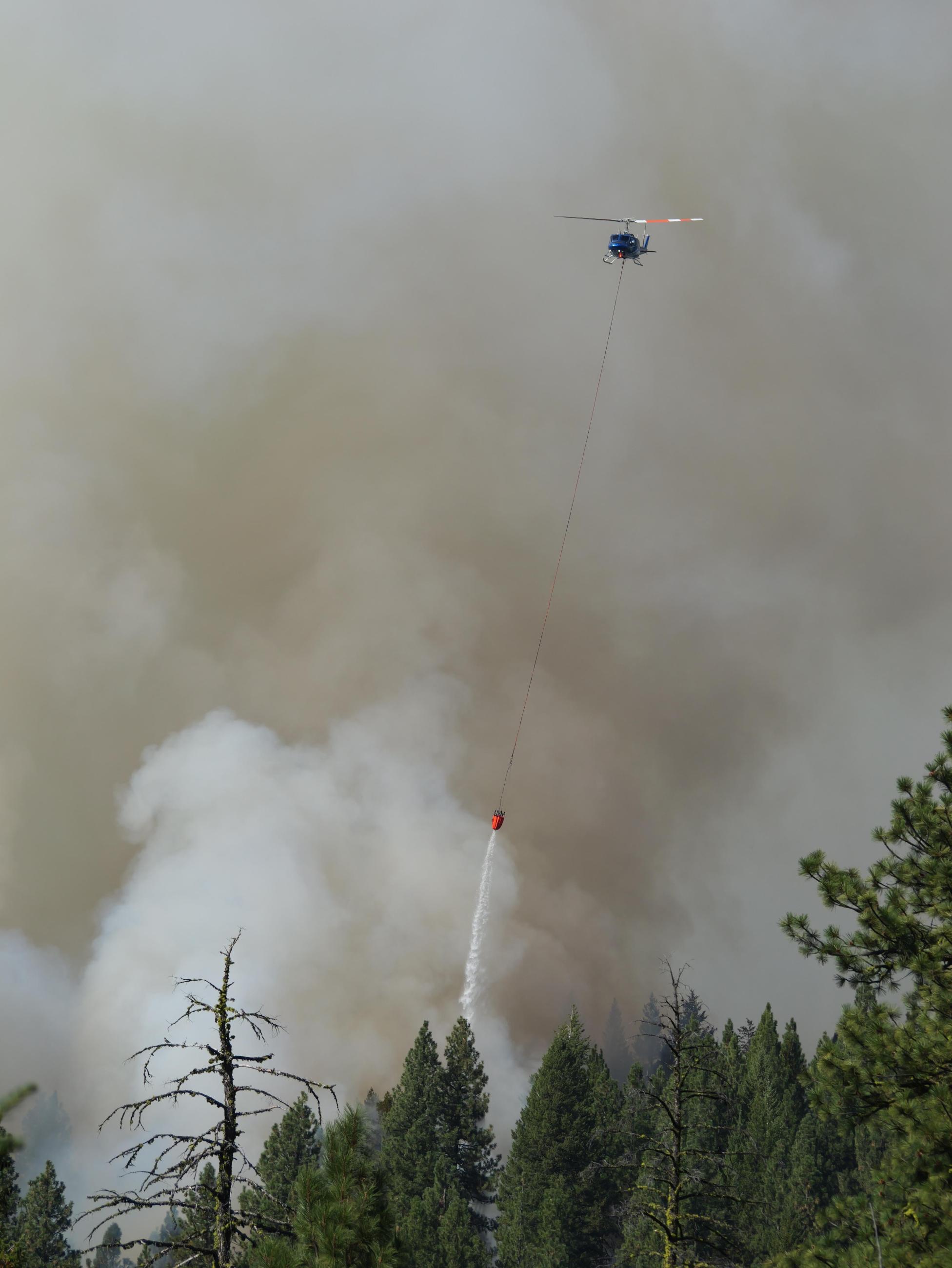 A helicopter with a water bucket attached is over a smoking forest as it drops the water.