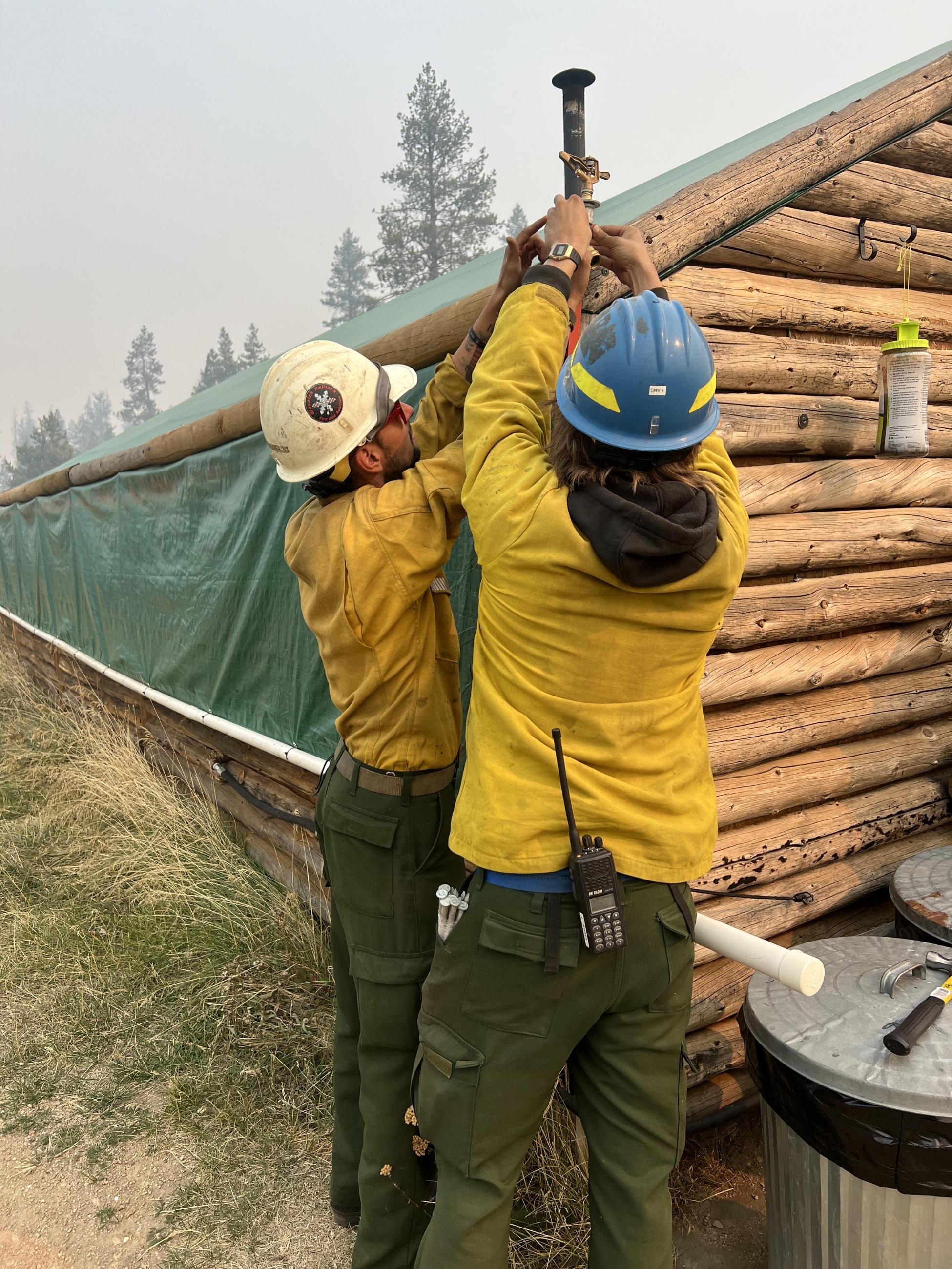 Two people wearing hard hats and yellow shirts place a sprinkler on the corner of a log cabin.