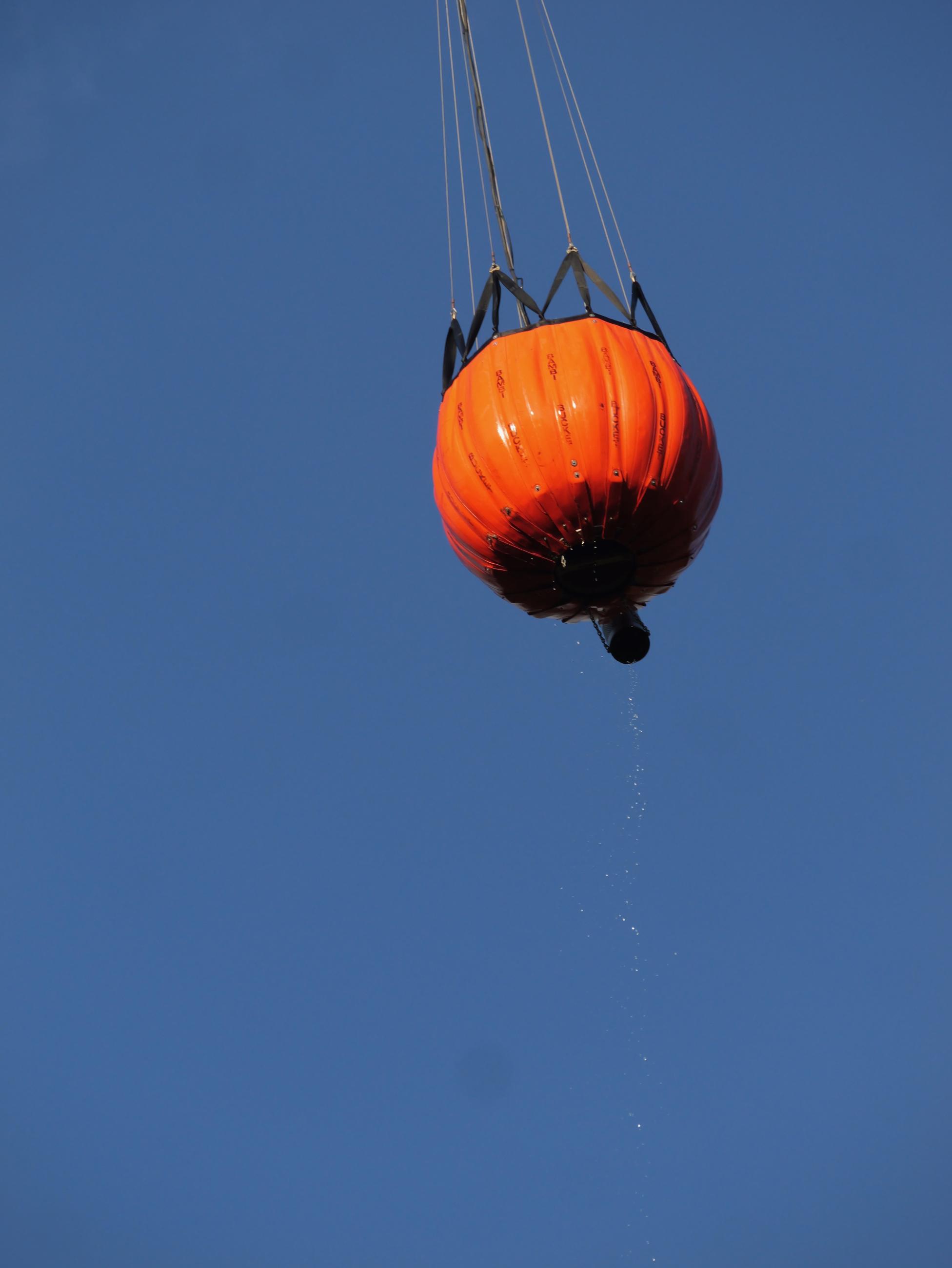 A bulging orange water bucket is suspended against a blue sky. Water drops off the bucket.