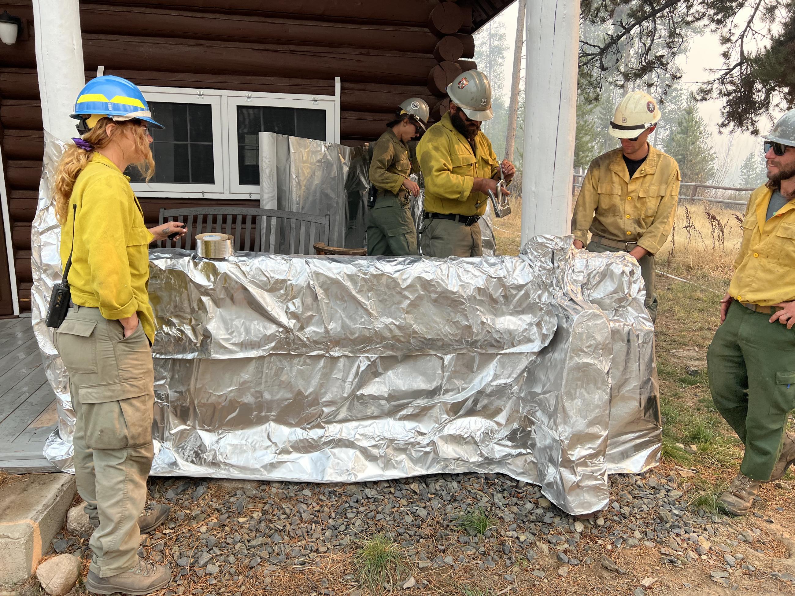 Firefighters in yellow shirts and helmets are seen installing fire-resistant foil wrapping on the porch of an historic cabin.