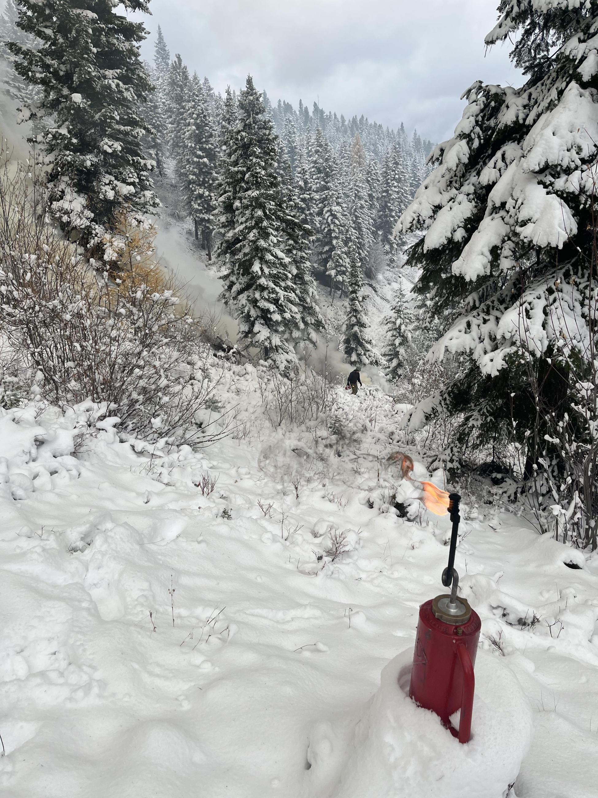 Drip torch sitting on the ground with snow and a pile burning in the background