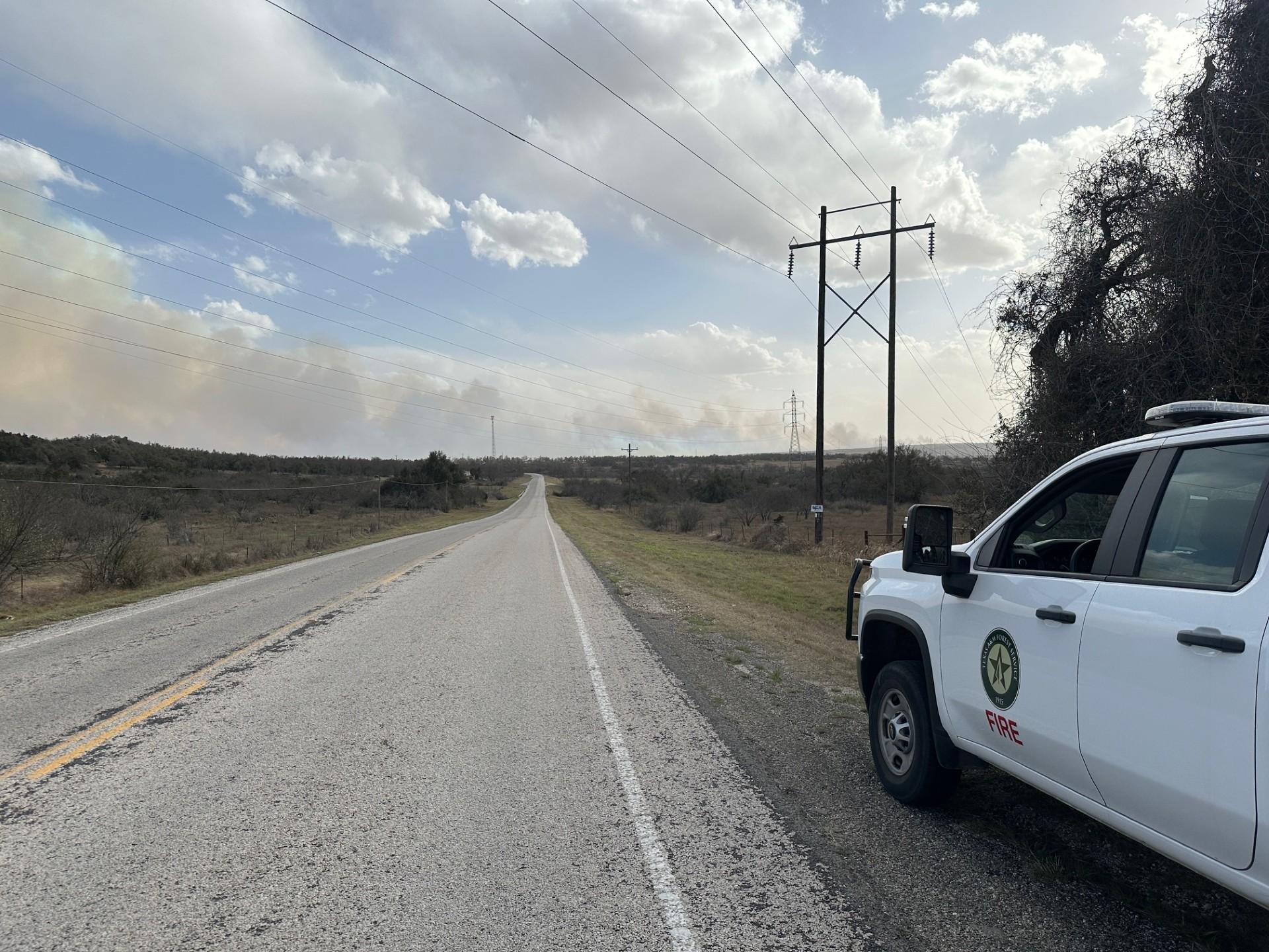 Photo shows a road in the middle, going up towards a blue sky with white, fluffy clouds. On the lower right, a Texas A&M Forest Service truck is parked and along the left, smoke lines the roadway.