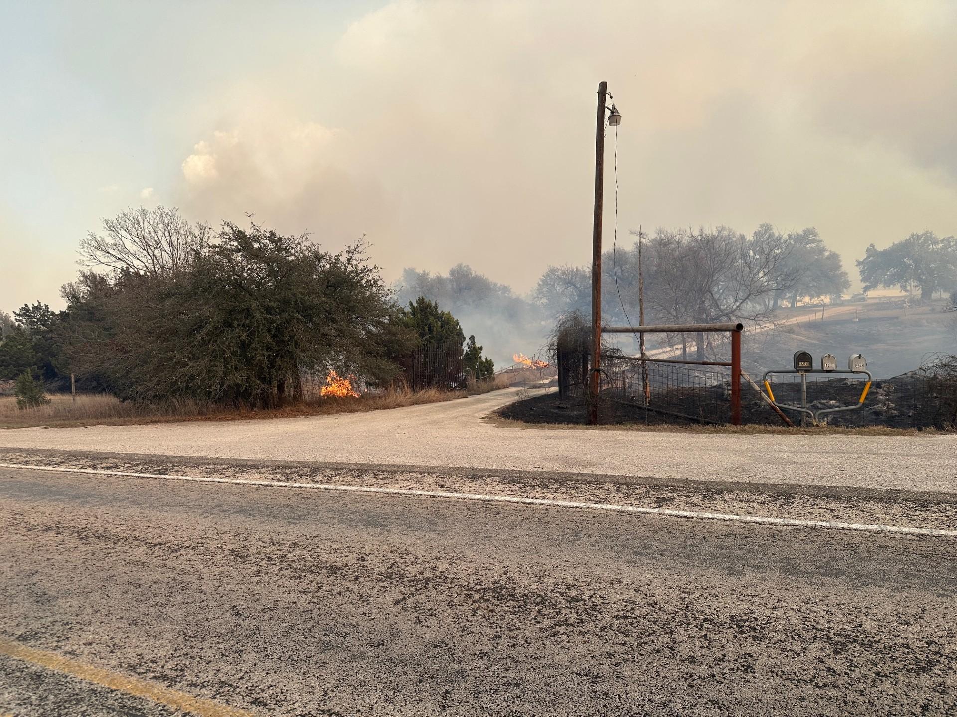 Photo of the fire with a utility pole in the center, surrounded by fluffy white smoke and a road in the front of the photo.