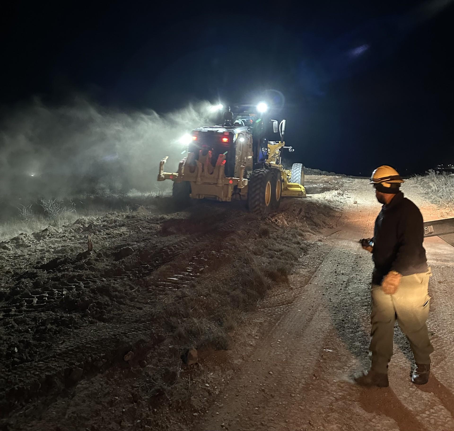 It is night time. A Firefighter is guiding a road grader by walking near it. 