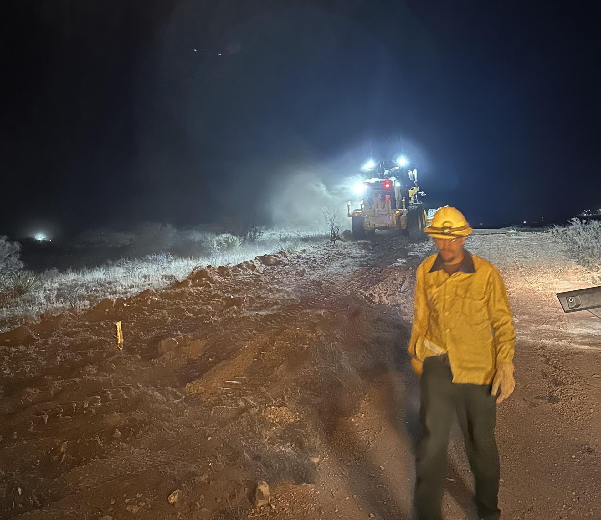 It is night. A firefighter is walking in front of a road grader that is improving containment line on the fire. 