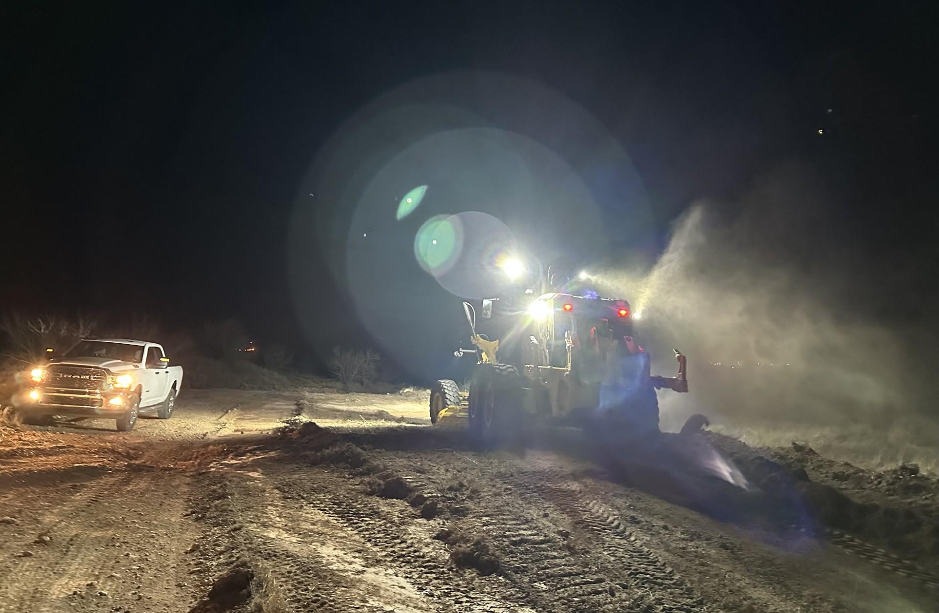 It is night. A road grader is working to improve fire containment line on the right of the photo and a white pickup truck is next to is on the left. 