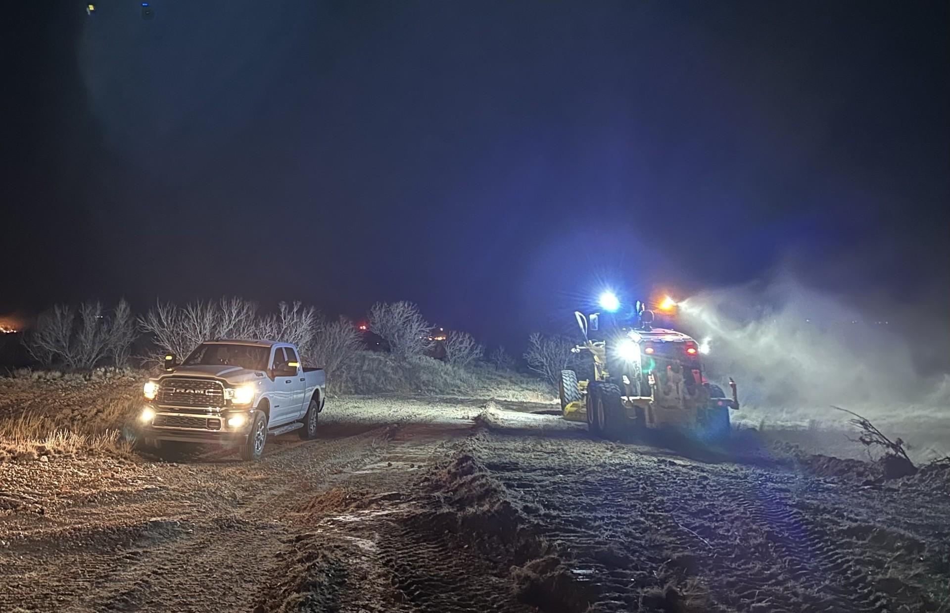 Nighttime operations show a pick up truck on the left and a road grader on the right, working to improve fire containment lines. 