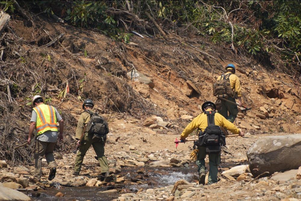 Four people in PPE cross stream near heavy storm debris