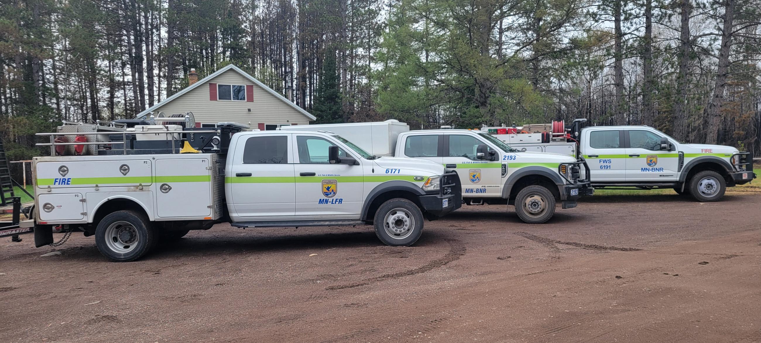 A strike team of three U.S. Fish & Wildlife Service utility wildland fire vehicles parked in front of a residence, positioned for structure protection.