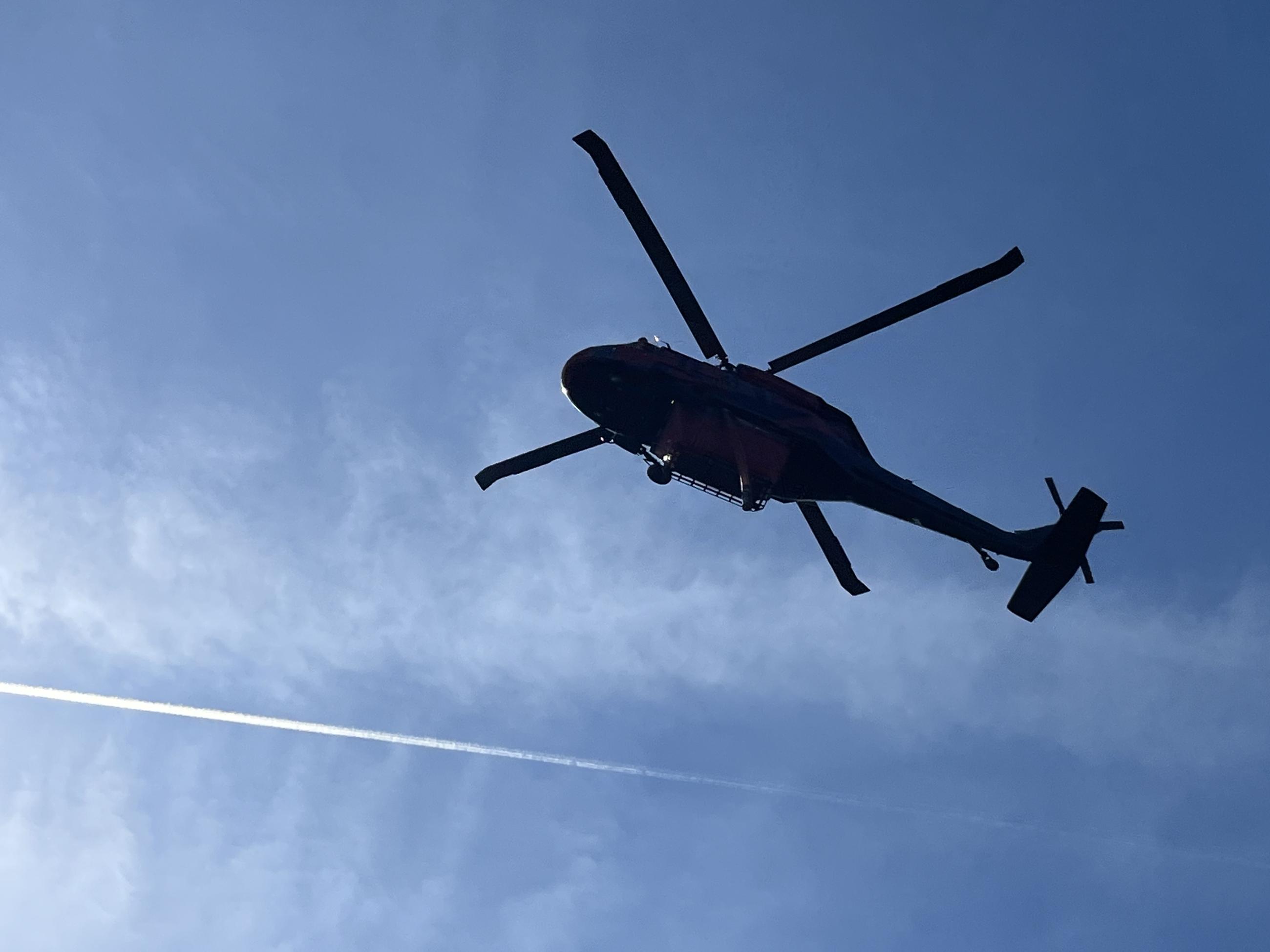 Helicopter silhoutetted against a bright blue sky as it flies overhead.