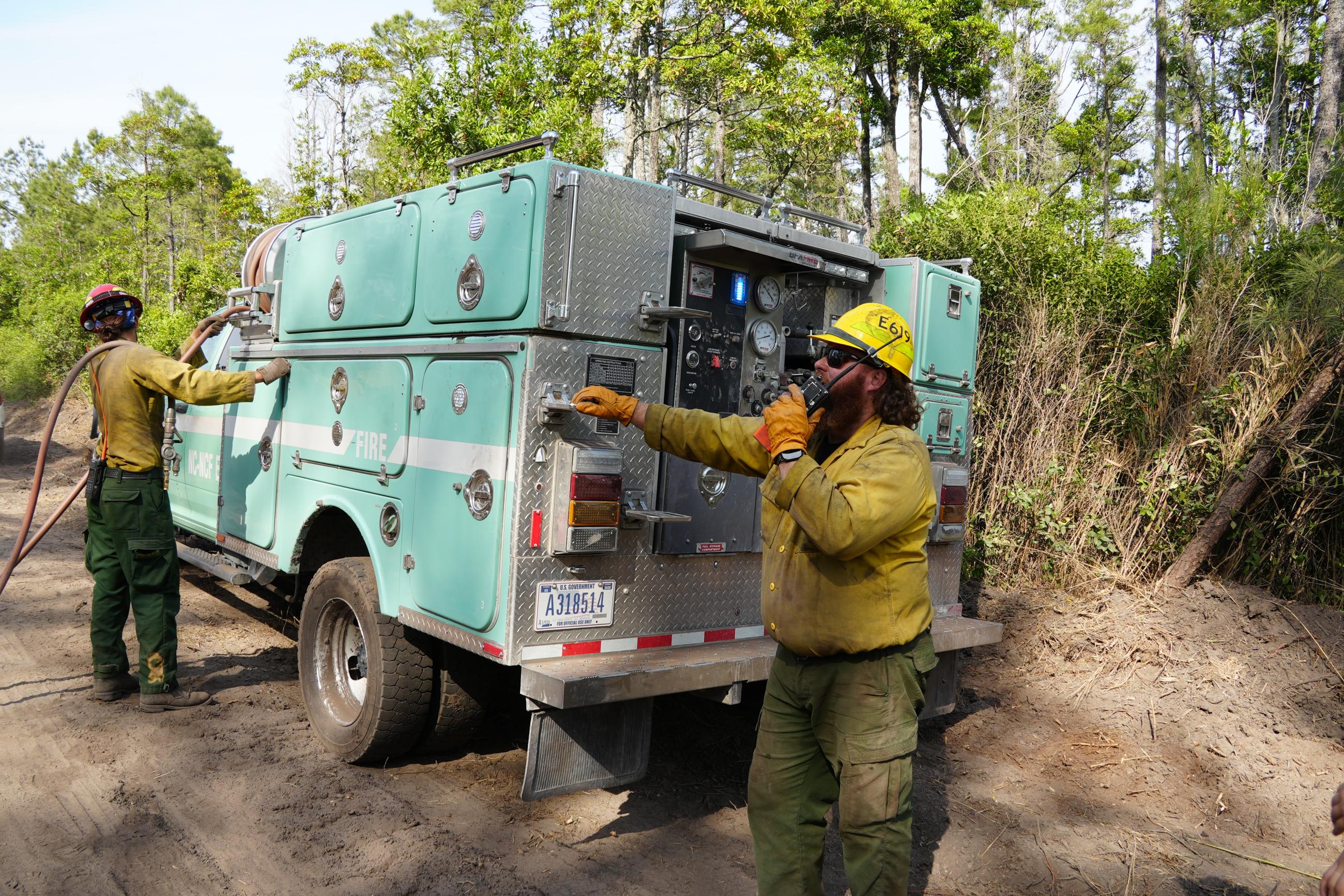 Firefighters standing next to engine. One is using the radio, the other is putting away the hose 
