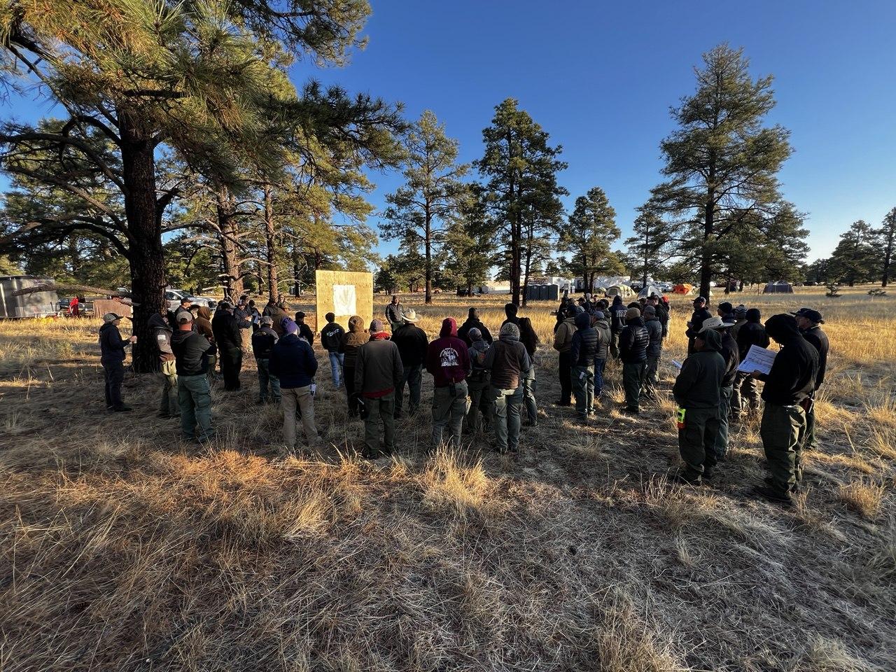 dozens of people stand in a circle near a large board with a map on it.