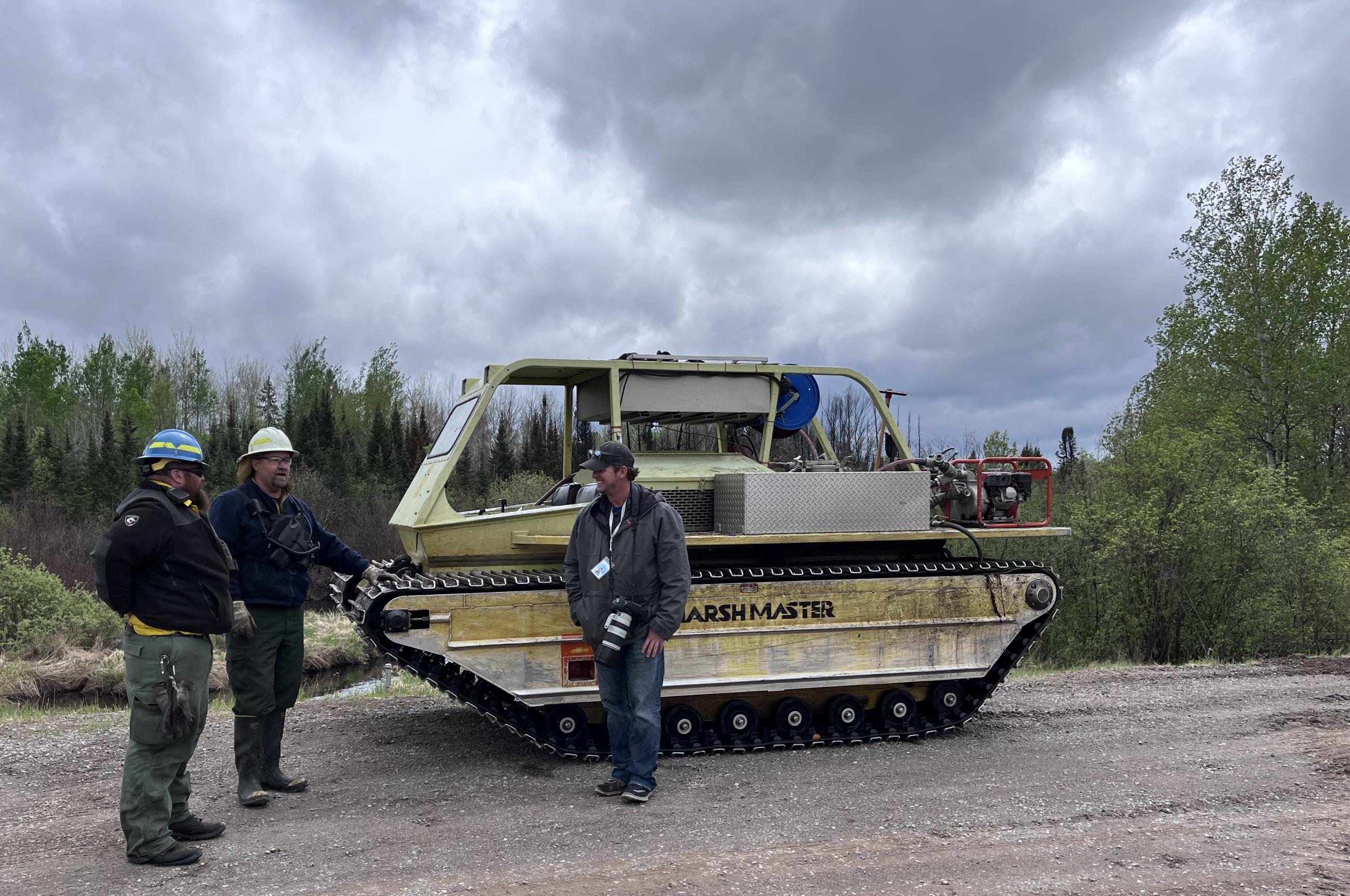 Two crew members and a reporter standing next to an amphibious tracked vehicle used to access wet areas