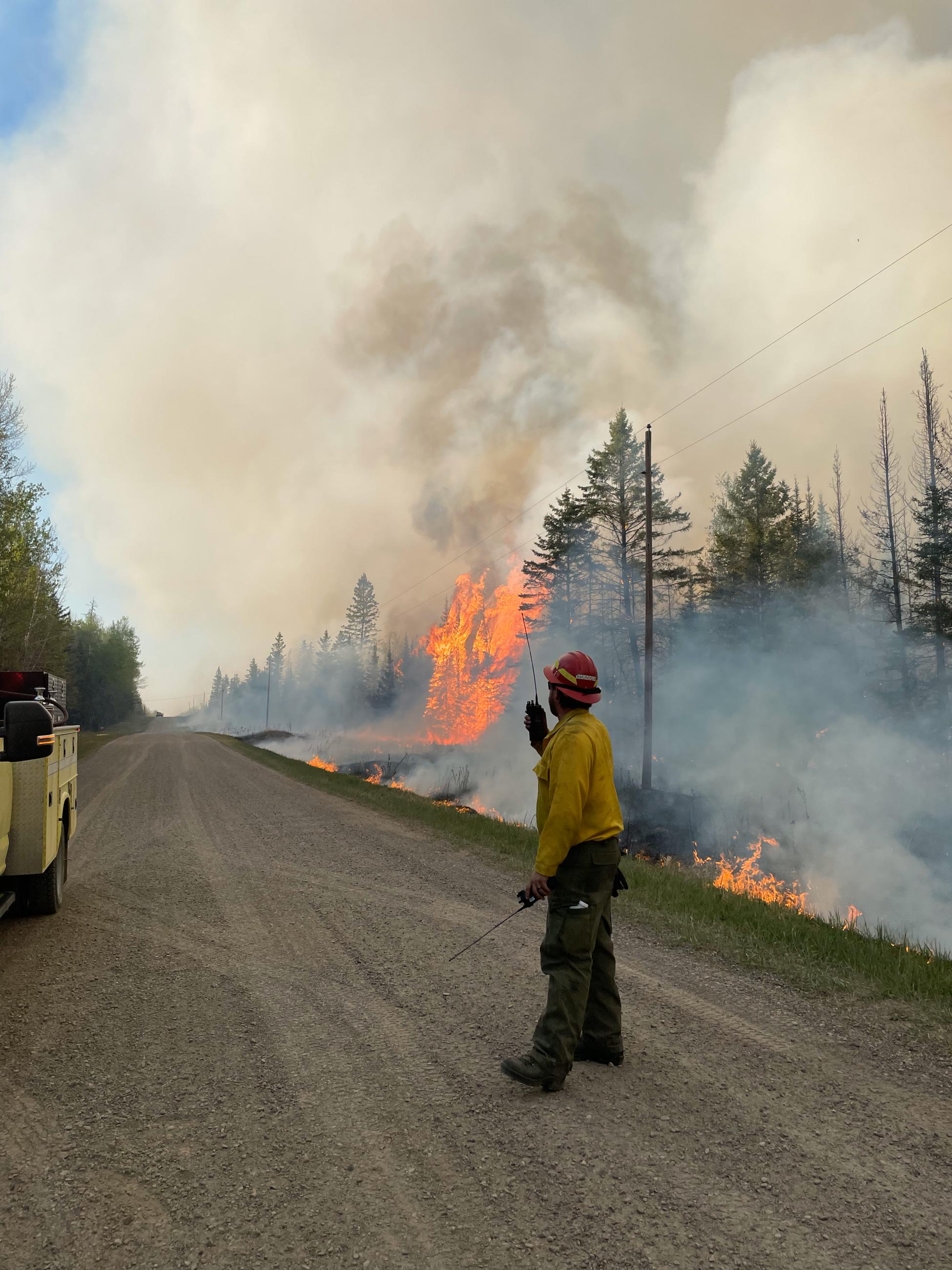 Image of a firefighter standing on a road near a yellow truck with a fire scene in the background