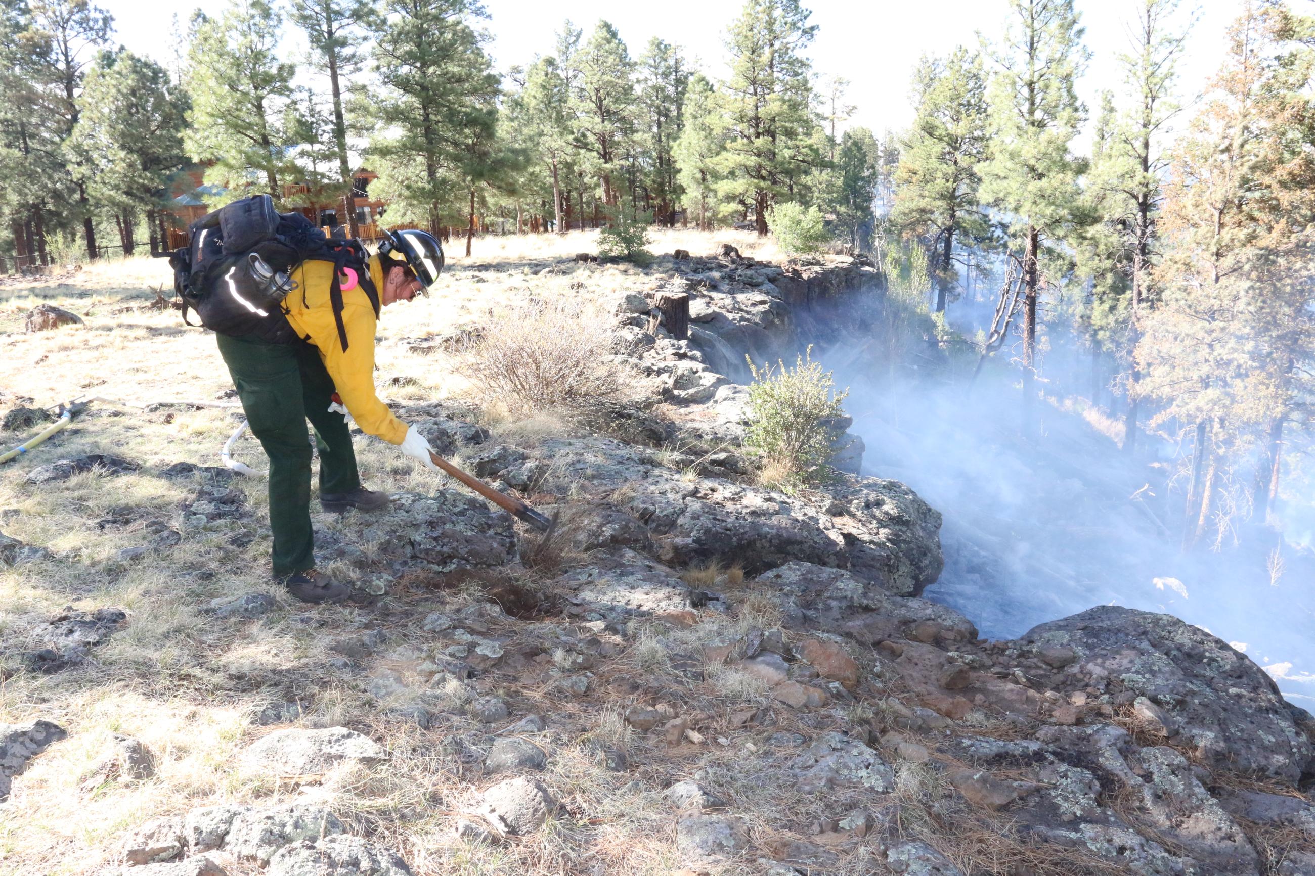 Person in yellow shirt with pack and tool digs in the dirt as smoke rises from the ground. 