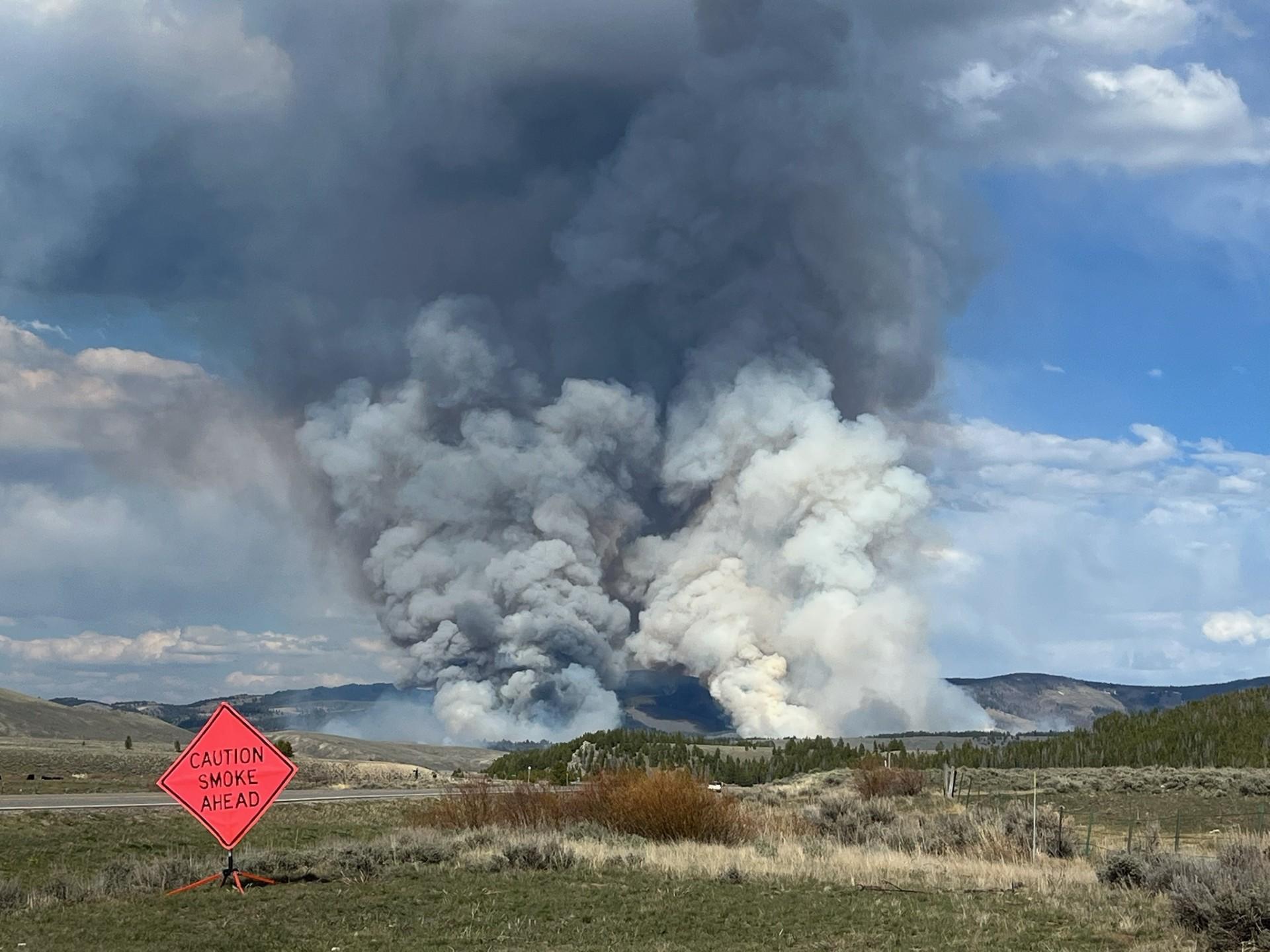 Two columns rise from the Sawlog fire, with a caution smoke ahead sign in the foreground along the highway.