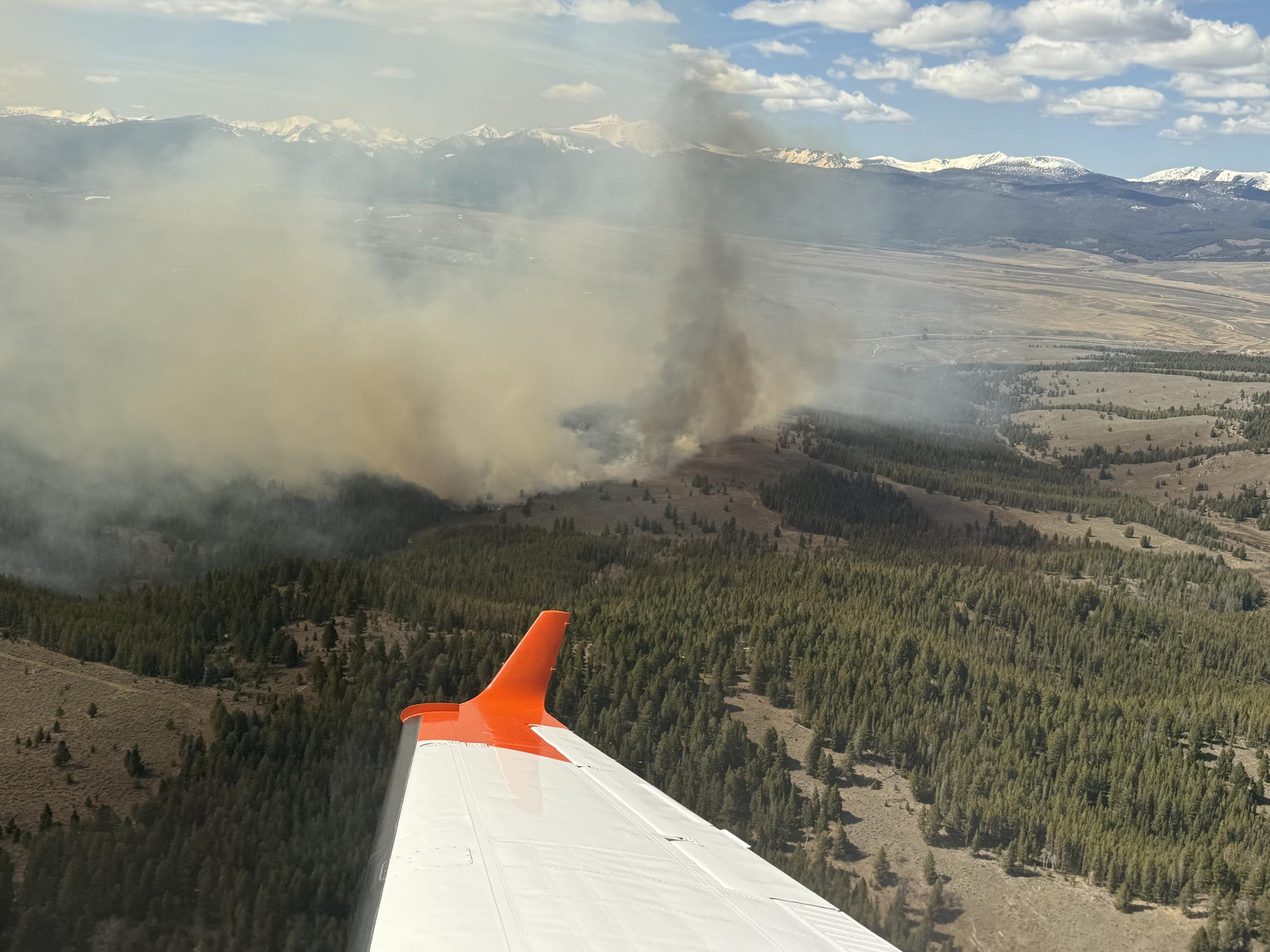 Smoke column rises from the timbered forest, with a wing from the lead plane visible in the foreground.