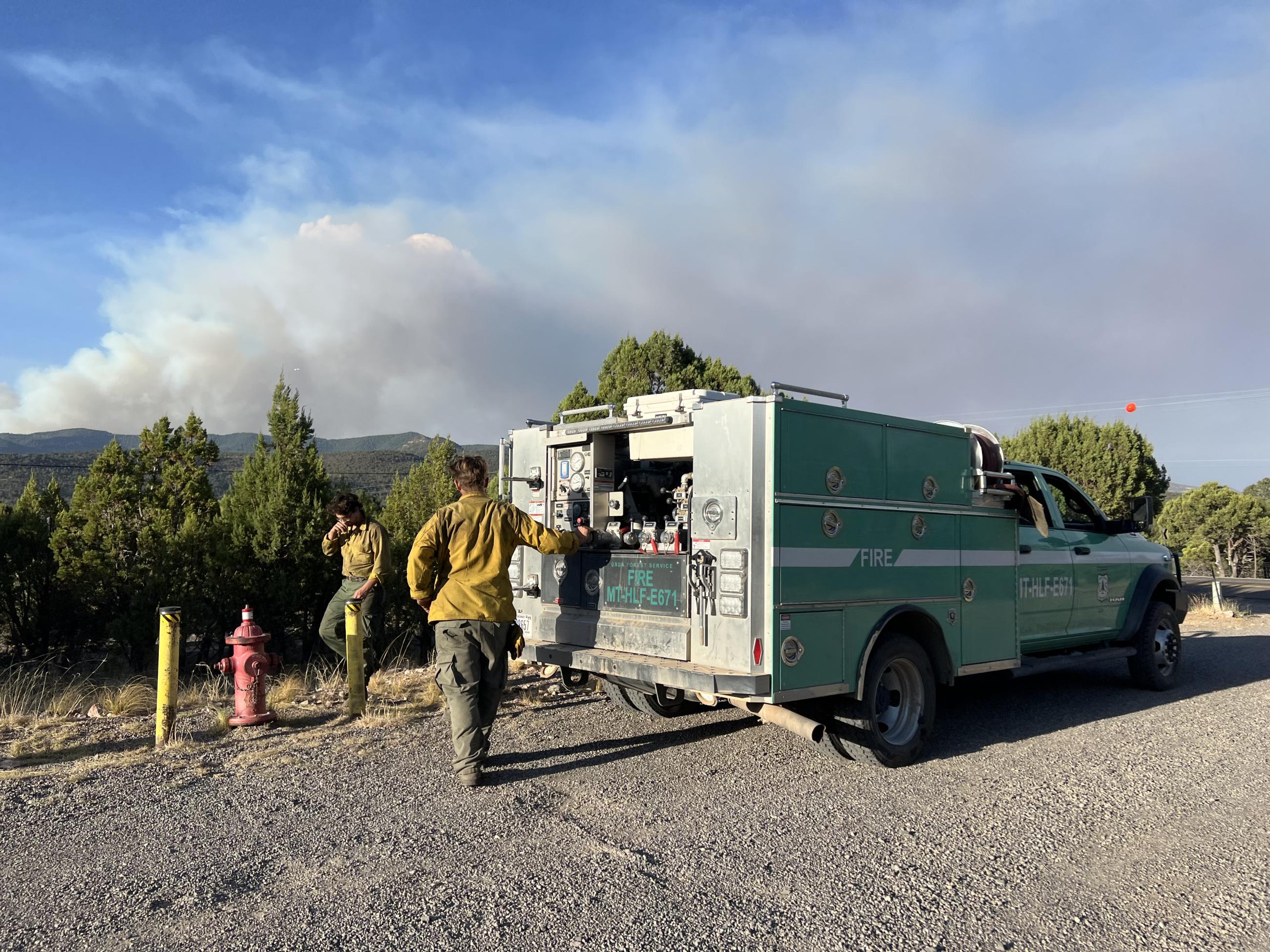 A mint green fire fighting apparatus is parked near a red fire hydrant. Workers in green pants and yellow shirts walk with tools to begin refilling the truck with water